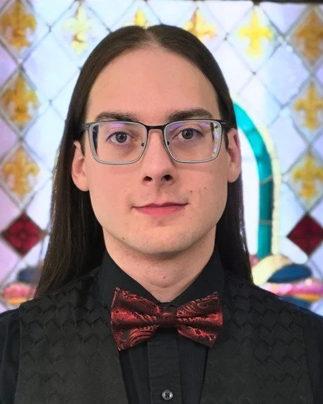 Daniel Pearsall, Bass. A young man with long dark hair, wearing glasses, a black shirt, and a red bow tie, standing in front of a decorative background with stained glass and floral patterns.