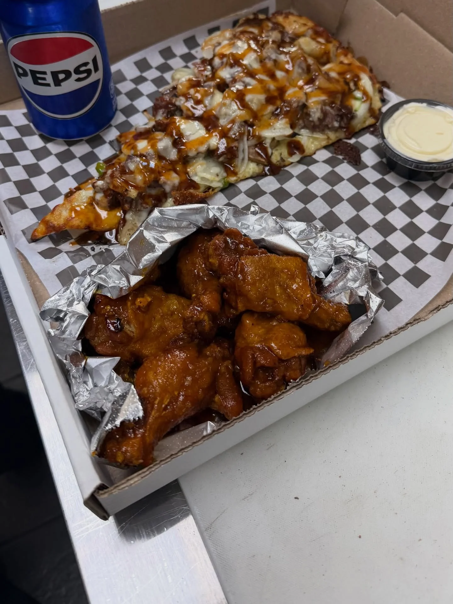 A box of fried chicken pieces, a slice of pizza with toppings, a small container of dipping sauce, and a can of Pepsi on checkered paper inside a pizza box.