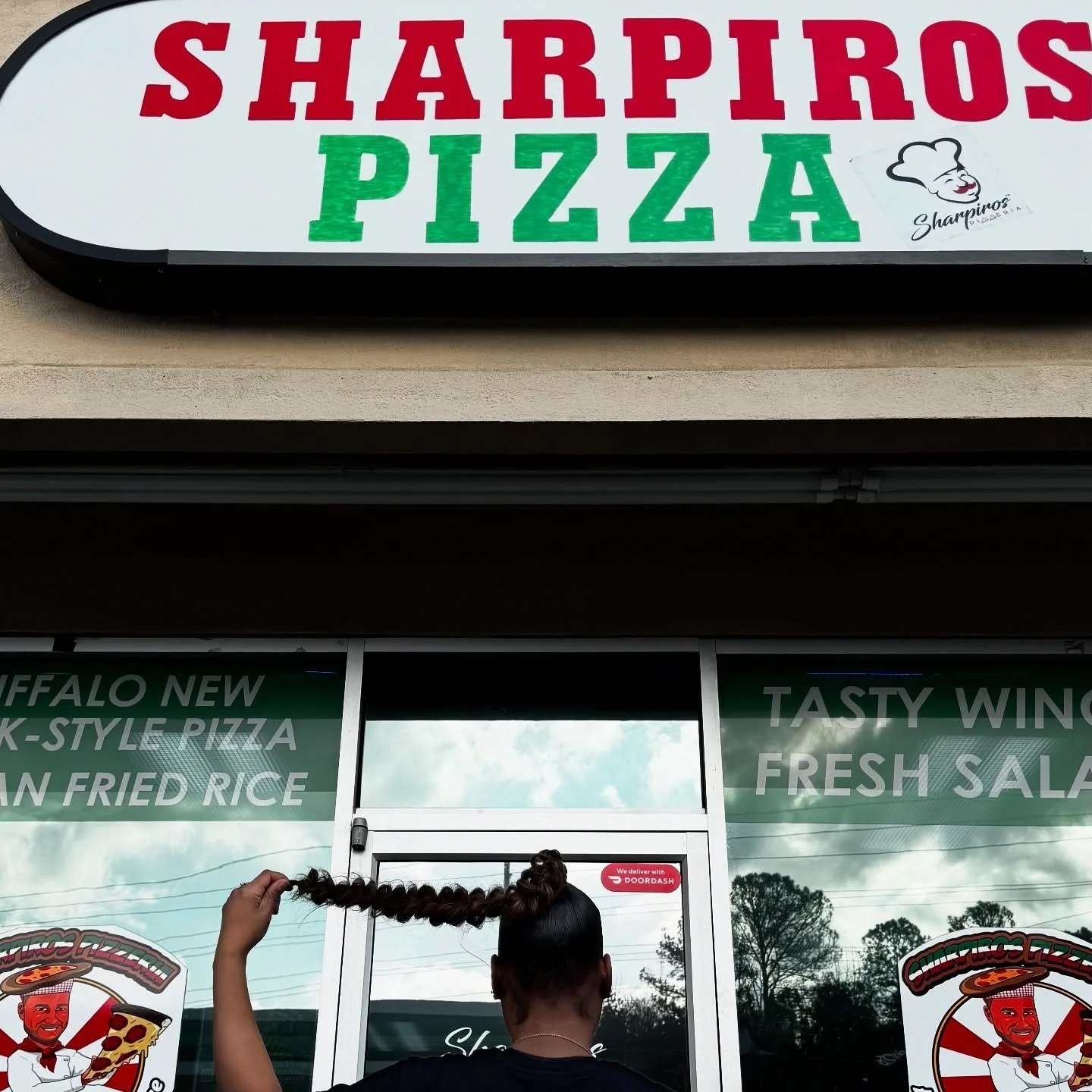 Sign for Sharpiro's Pizza above the entrance of a pizza restaurant, with a logo of a smiling chef. A person with dark hair tied in a bun is holding or playing with their braided hair in the foreground. The establishment offers phone ordering and delivery through DoorDash.