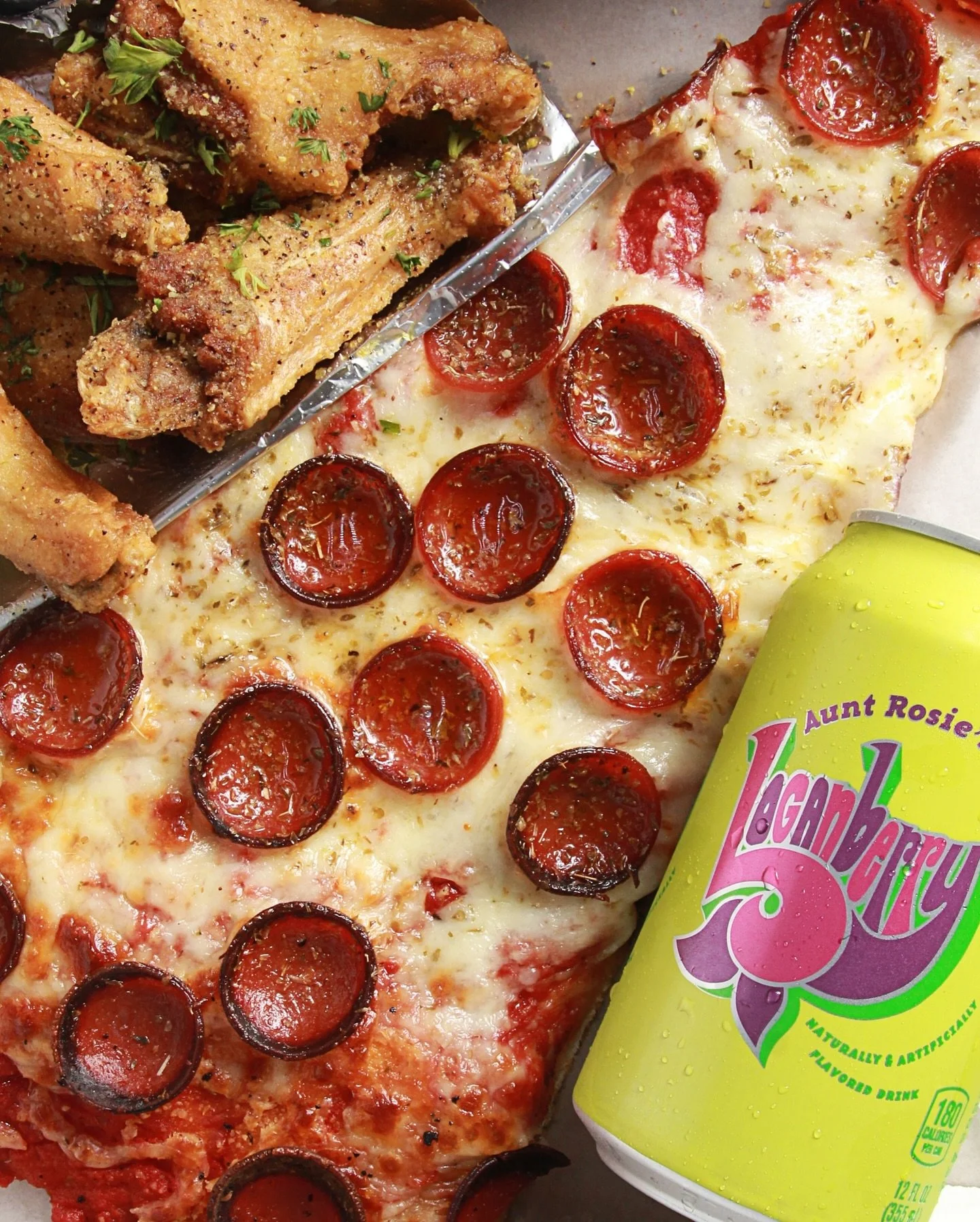 A close-up of a slice of pepperoni pizza with melted cheese, a side of fried chicken pieces garnished with parsley, and a can of grape-flavored drink with condensation on the surface.