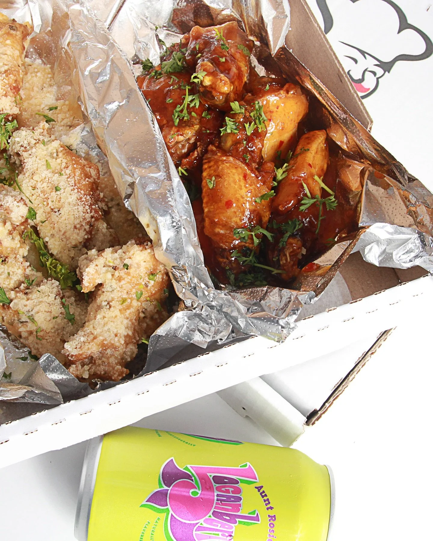 Fried chicken pieces with breadcrumbs garnish on the left, and a container of chicken drumsticks with barbecue sauce and parsley on the right, inside a cardboard box lined with aluminum foil. A can of fruit punch juice is below the box.