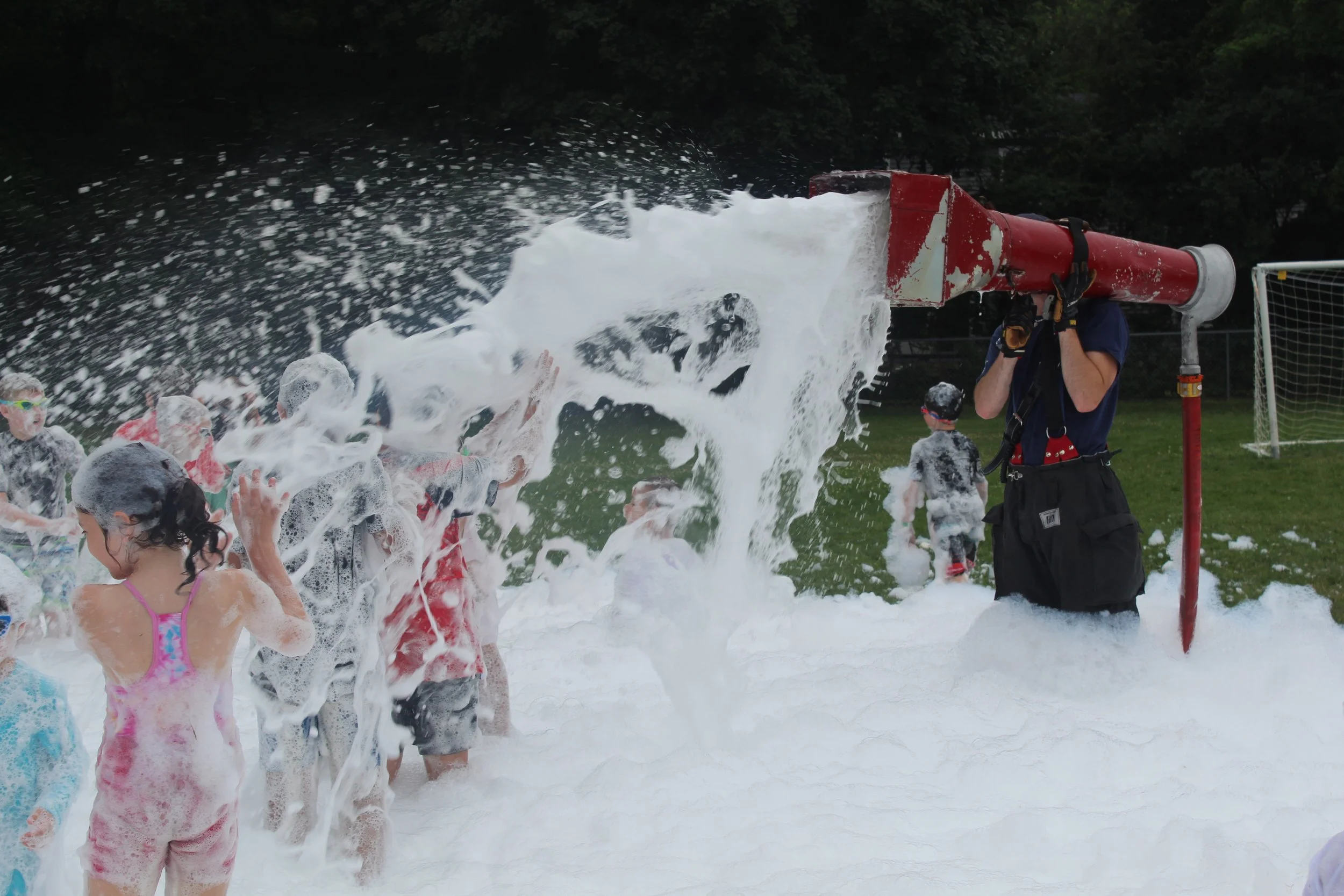 Children playing in foam with a person operating a foam machine outdoors.