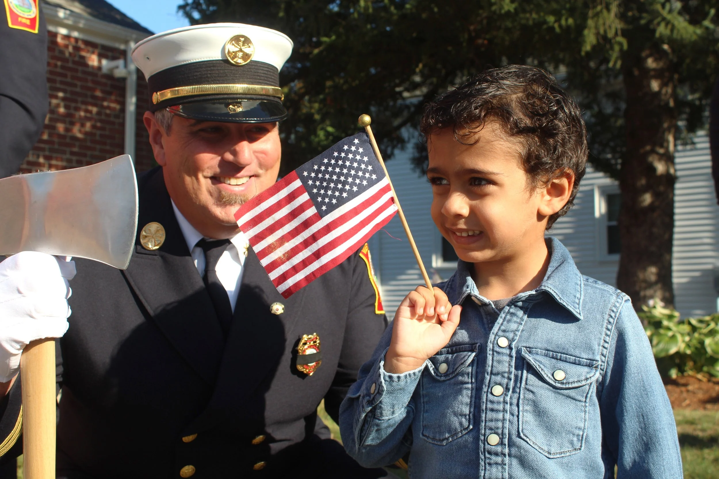 A firefighter in dress uniform and a young boy holding an American flag are smiling together during a patriotic event outdoors.