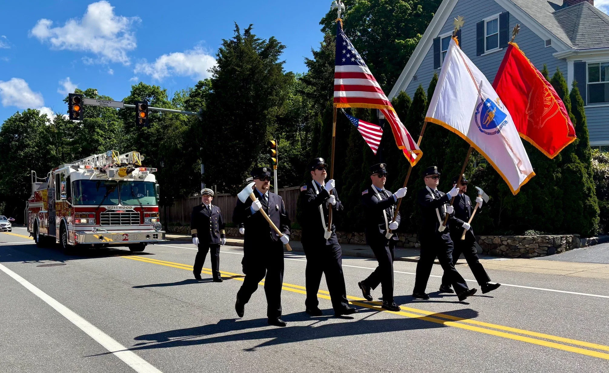 A parade with firefighters and honor guard carrying flags, including the American flag, on a street under a blue sky with clouds.
