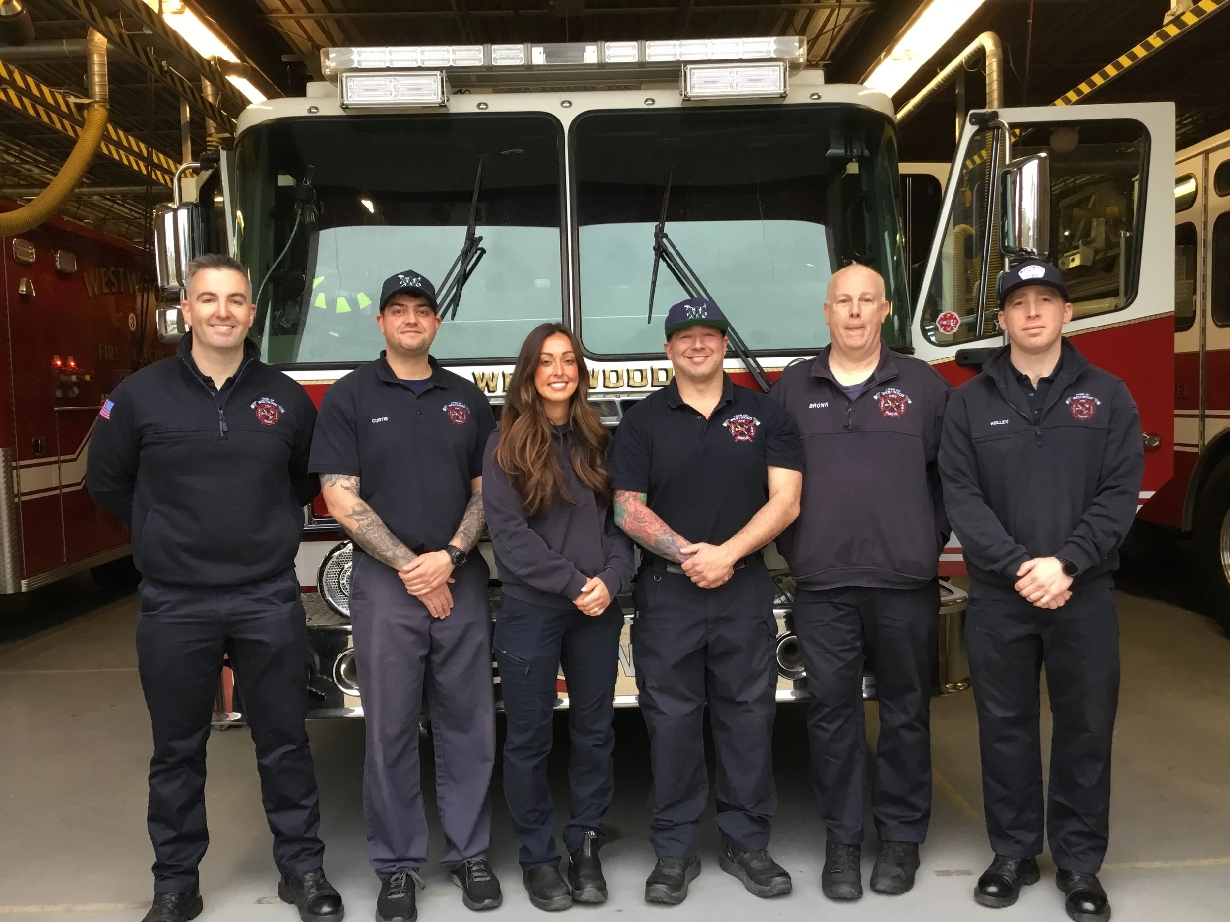 Group of six firefighters standing in front of a fire truck inside a fire station, smiling at the camera.