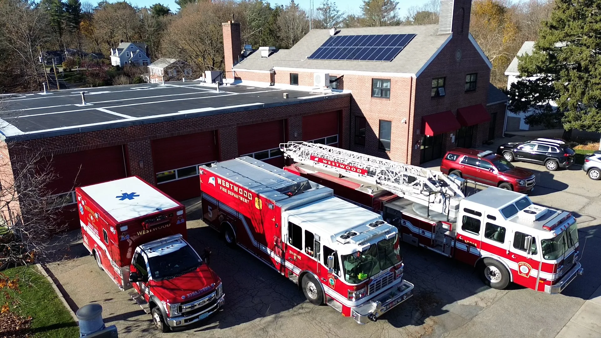Multiple red and white fire trucks parked outside a brick fire station with a garage door, alongside several parked cars. The fire trucks are labeled Westwood Fire Department and include fire engines and ladder trucks. The background shows a building