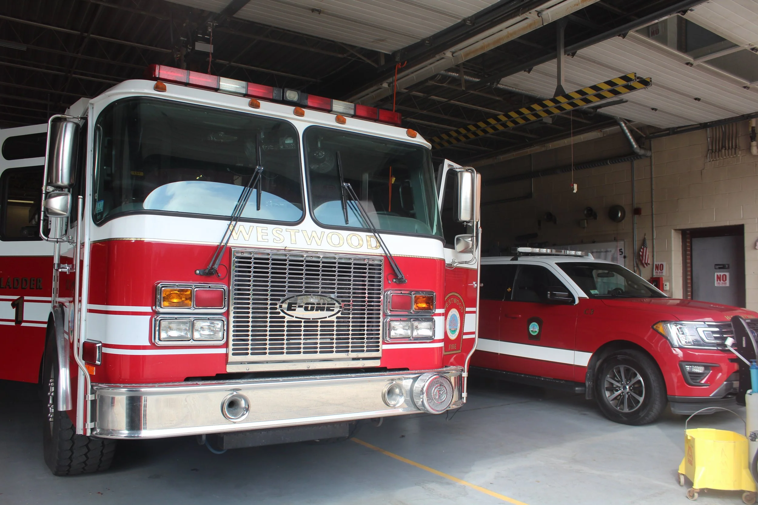 A fire truck labeled 'Westwood' parked inside a fire station garage alongside a red and black utility vehicle. The garage has an overhead door, some yellow and black safety striping, and various equipment.