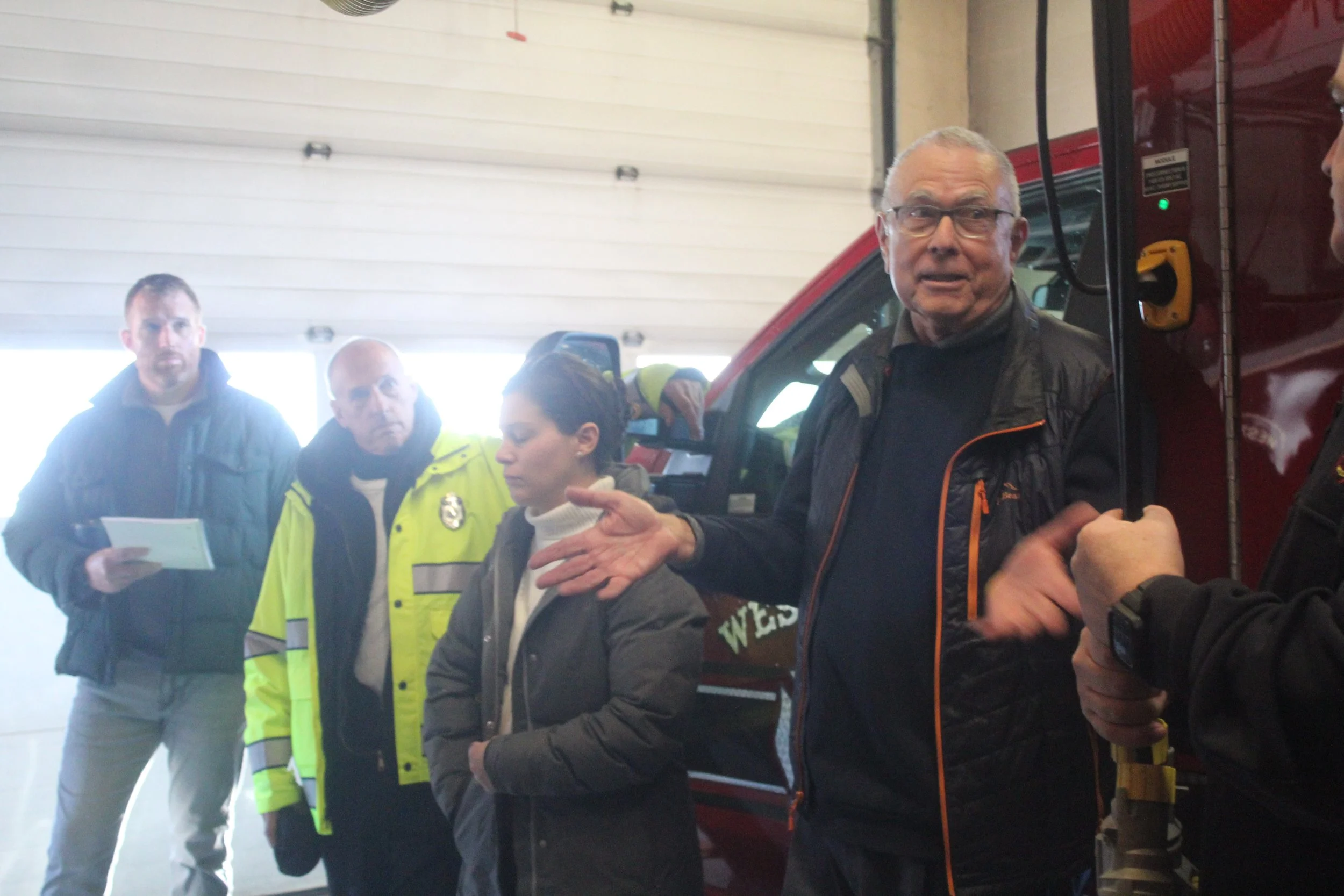 Group of five people, including firefighters and a man reading a document, gathered near a red fire truck inside a building with a raised garage door.