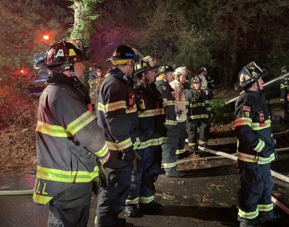 Group of firefighters standing in a line at night, wearing protective gear and helmets, with fire hoses on the ground, in a wooded area.
