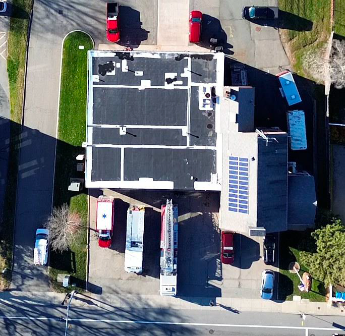 Aerial view of a building with a flat roof, surrounding parking lot with cars, and emergency vehicles including fire trucks parked nearby.