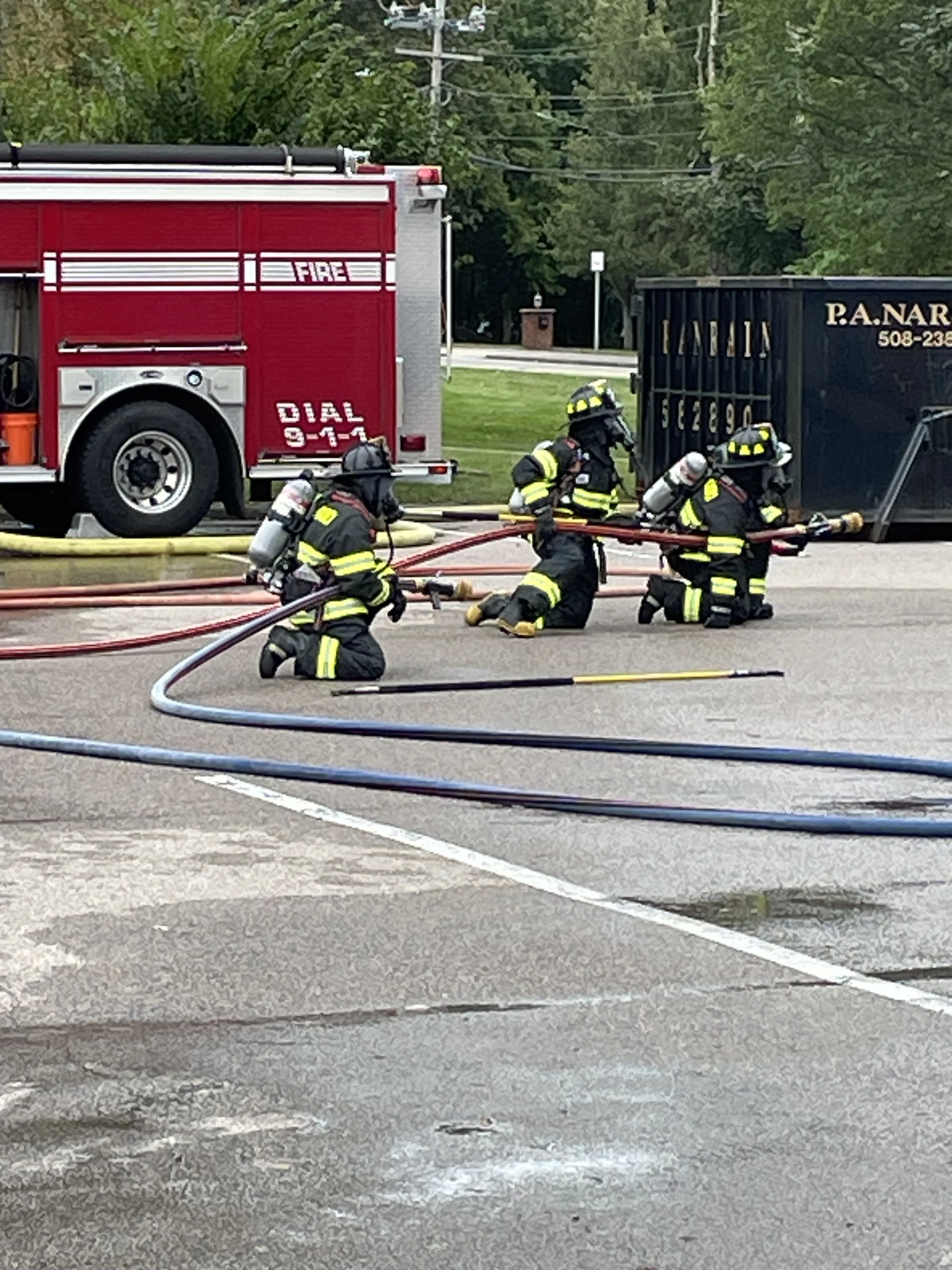 Four firefighters in full gear and oxygen tanks are kneeling and working on fire hoses on a paved street. A red fire truck and a black container with yellow markings are in the background, along with green trees and utility poles.