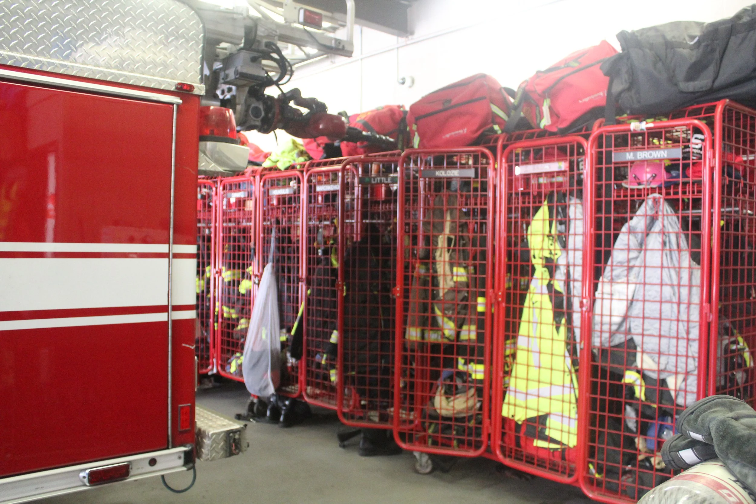 Firefighter gear on red carts at fire station, including helmets and protective clothing, with part of fire truck visible.