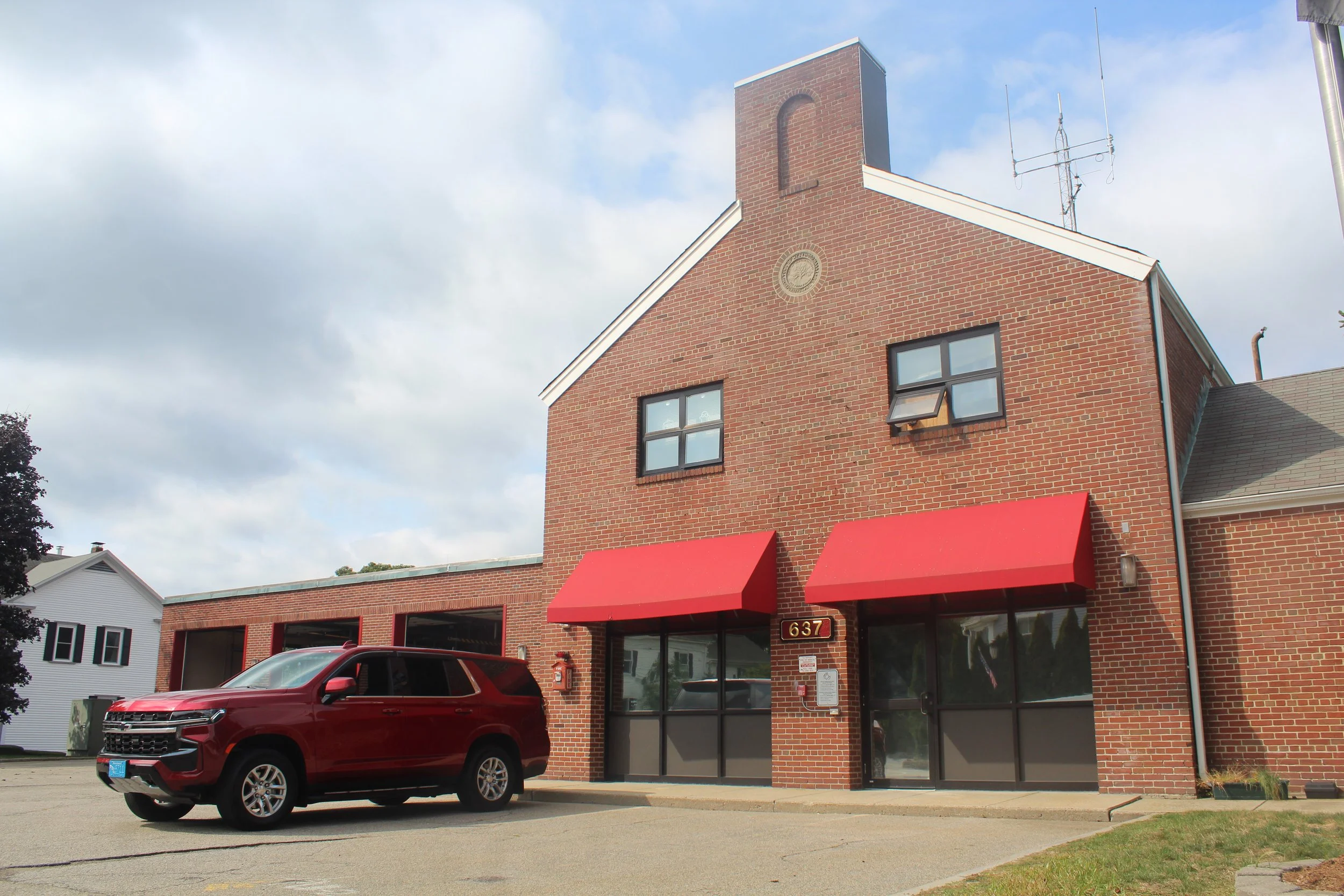 A red SUV parked in front of a brick building with red awnings and a glass door entrance, with a cloudy sky above.