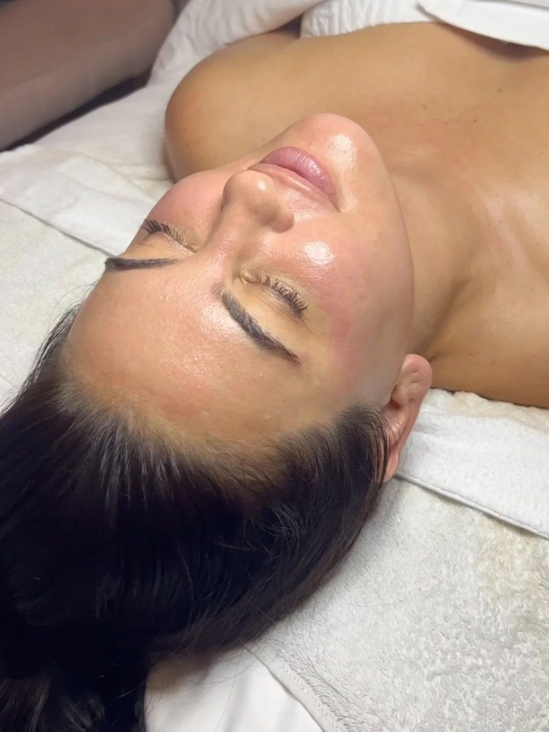 A woman lying on a bed with her eyes closed, receiving a facial treatment.