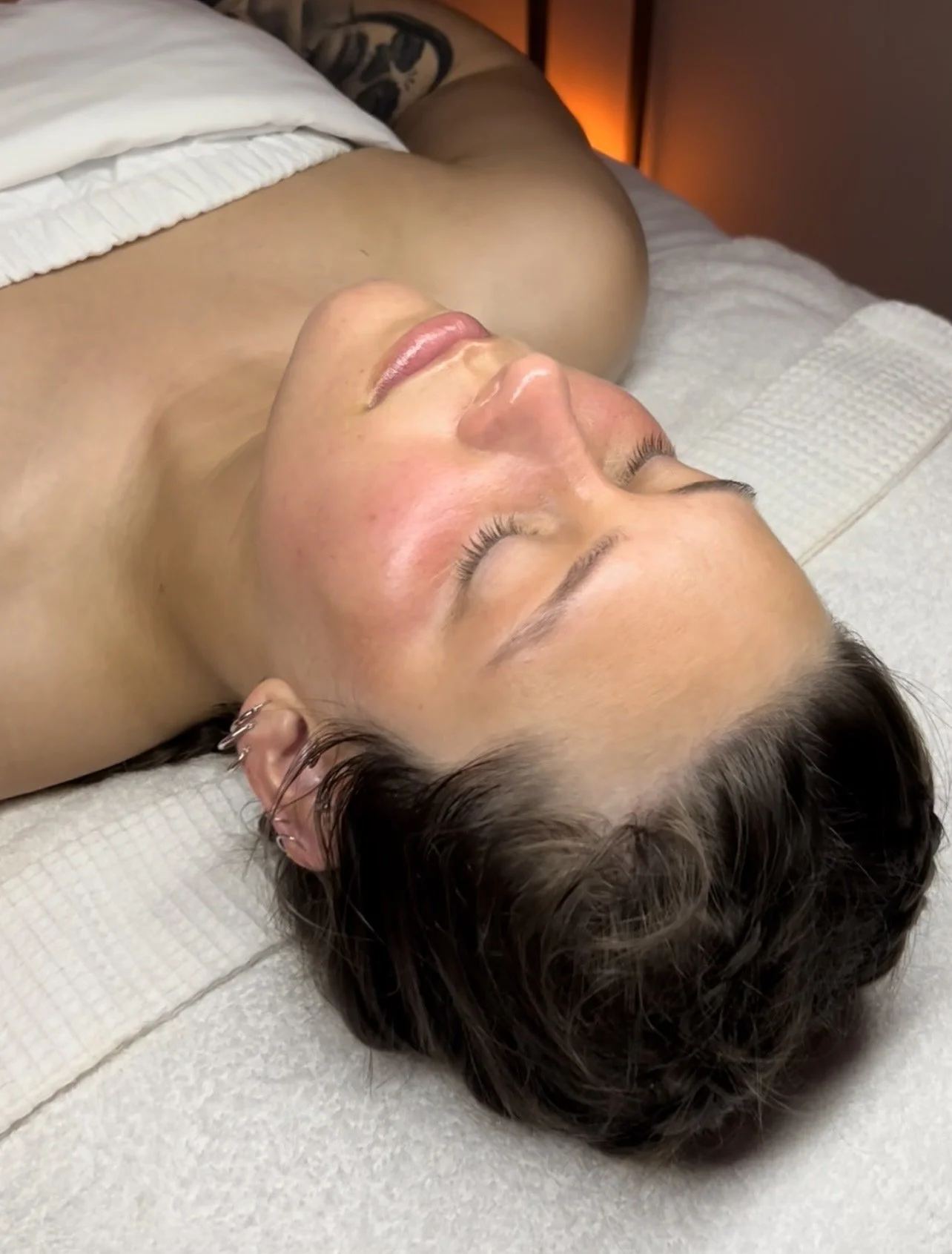 A woman lying on her back with eyes closed, appearing relaxed, positioned on a treatment table with a white towel beneath her head, in a softly lit room.