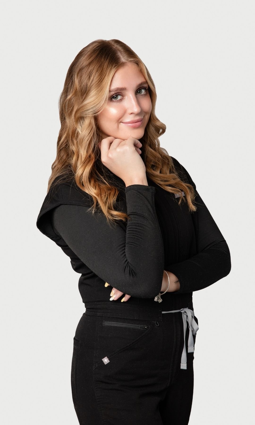 Young woman with long, wavy hair, wearing a black top and black pants, standing with her arms crossed and resting her chin on her right hand, smiling subtly, against a plain light background.
