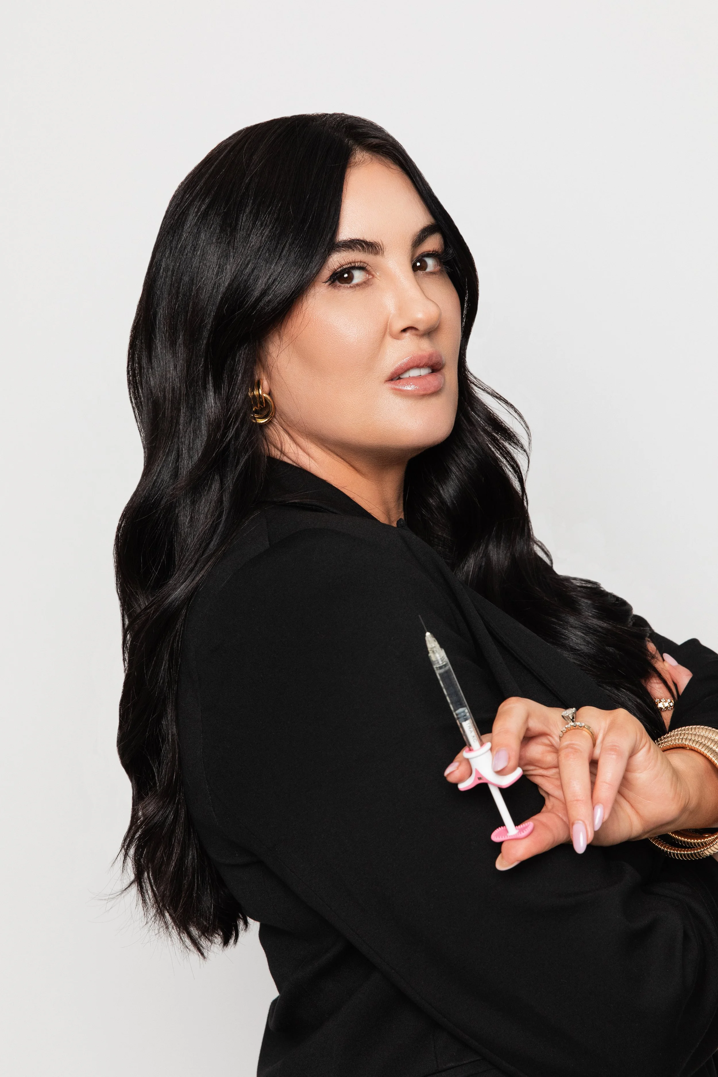 A woman with long black hair and gold jewelry holding a pink and white syringe on her arm, posing against a plain white background.