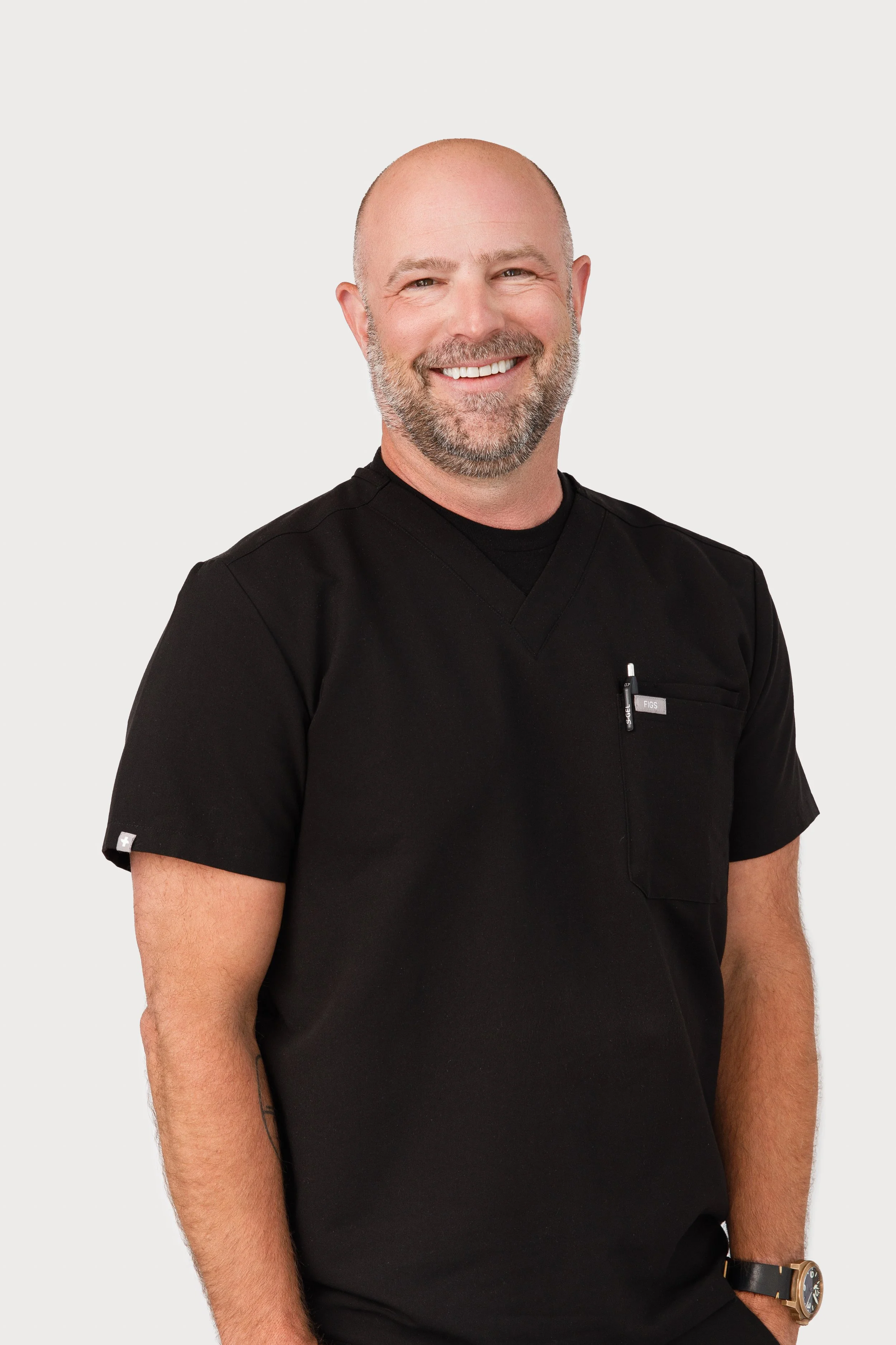 Smiling male healthcare professional wearing black scrubs and watch, standing against a plain light background.
