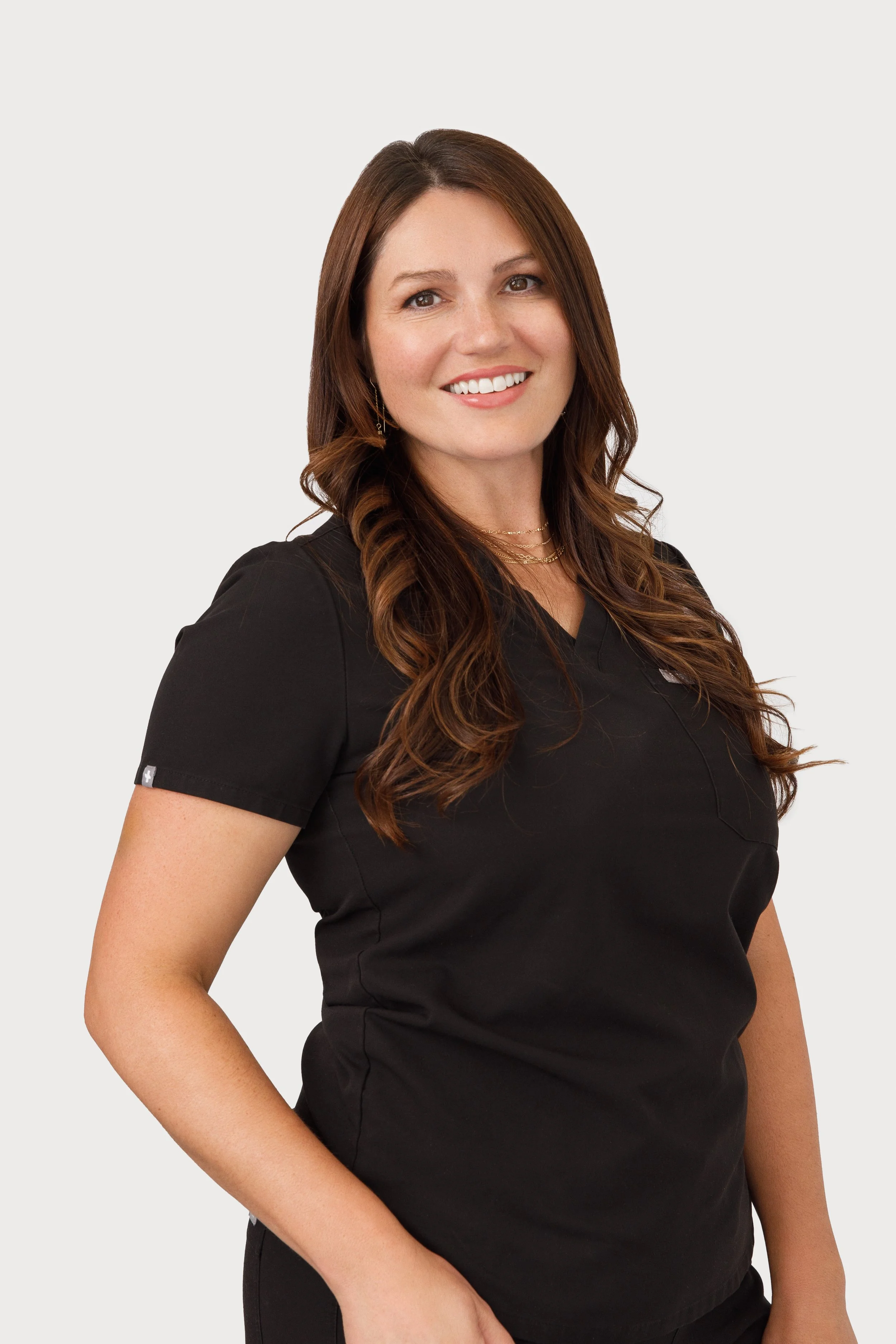A woman with long, wavy brown hair, wearing a black top, smiling with teeth showing, standing against a plain light background.