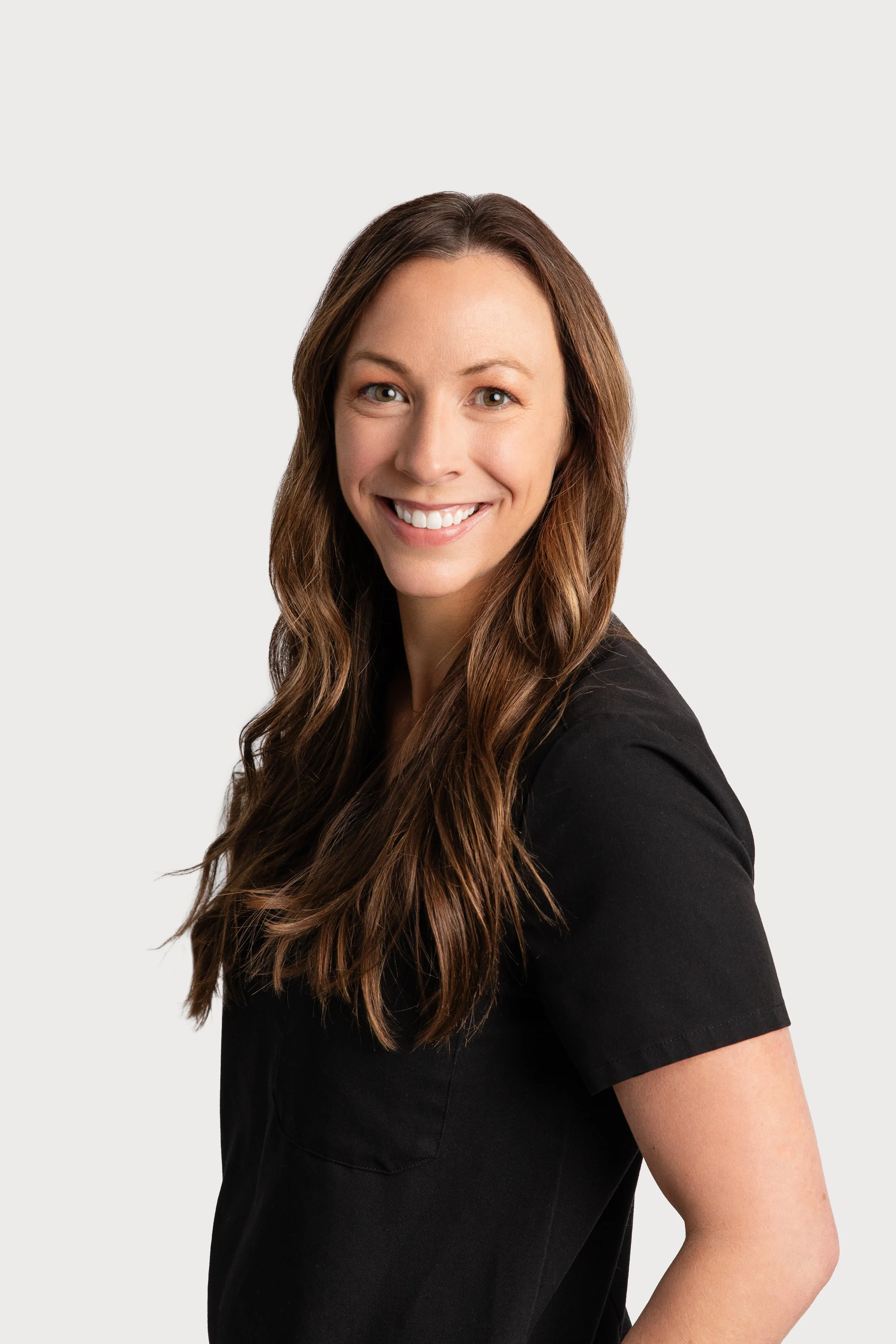 Portrait of a smiling woman with long brown hair, wearing a black top, against a plain light background.