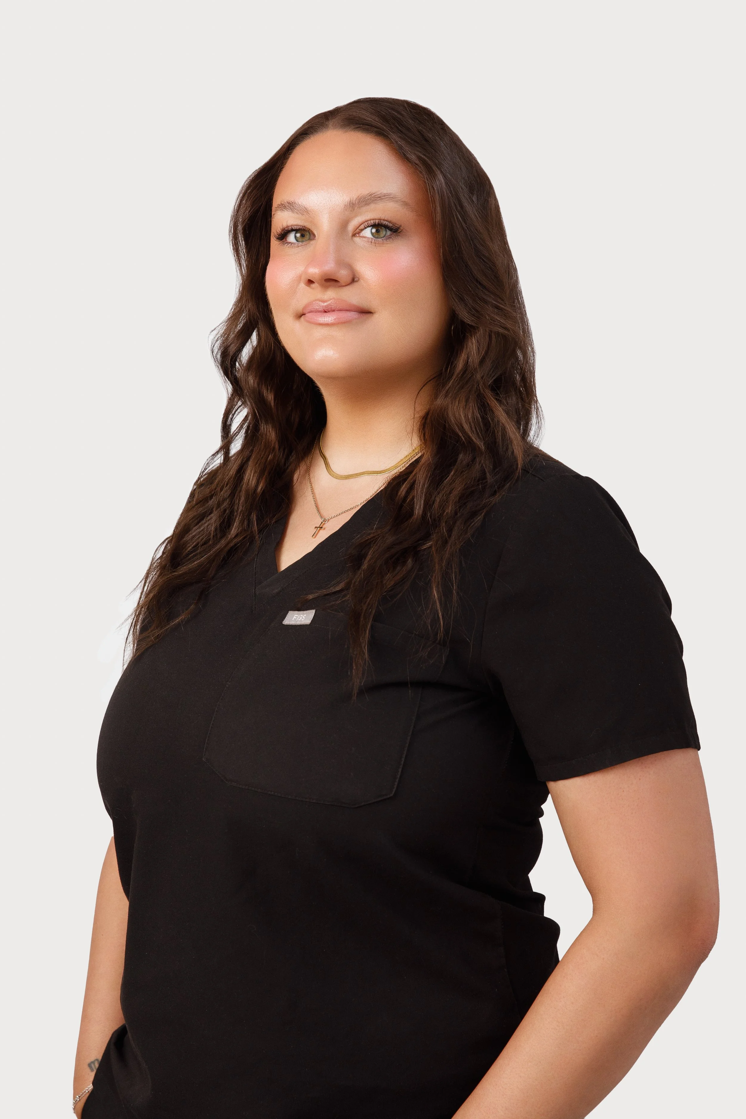 A woman wearing black scrubs, standing against a plain white background, with long wavy brown hair and a subtle smile.