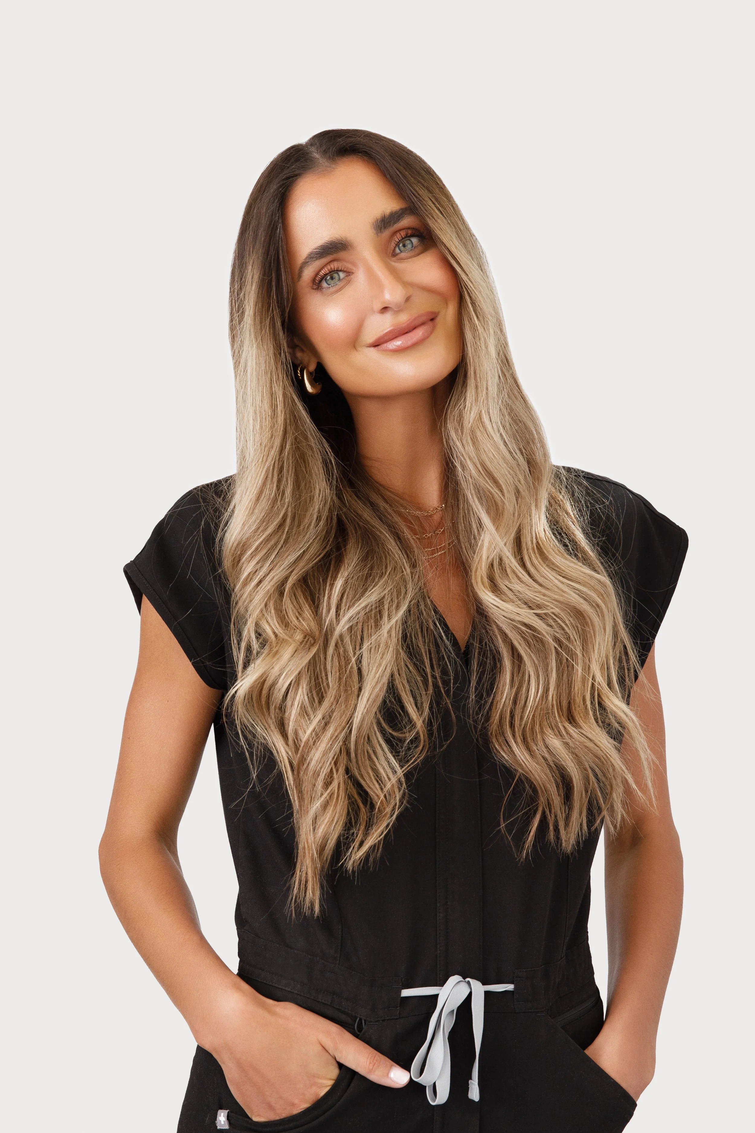 A woman with long wavy blonde hair wearing a black sleeveless top and gold hoop earrings, smiling with hands in her pockets, against a white background.
