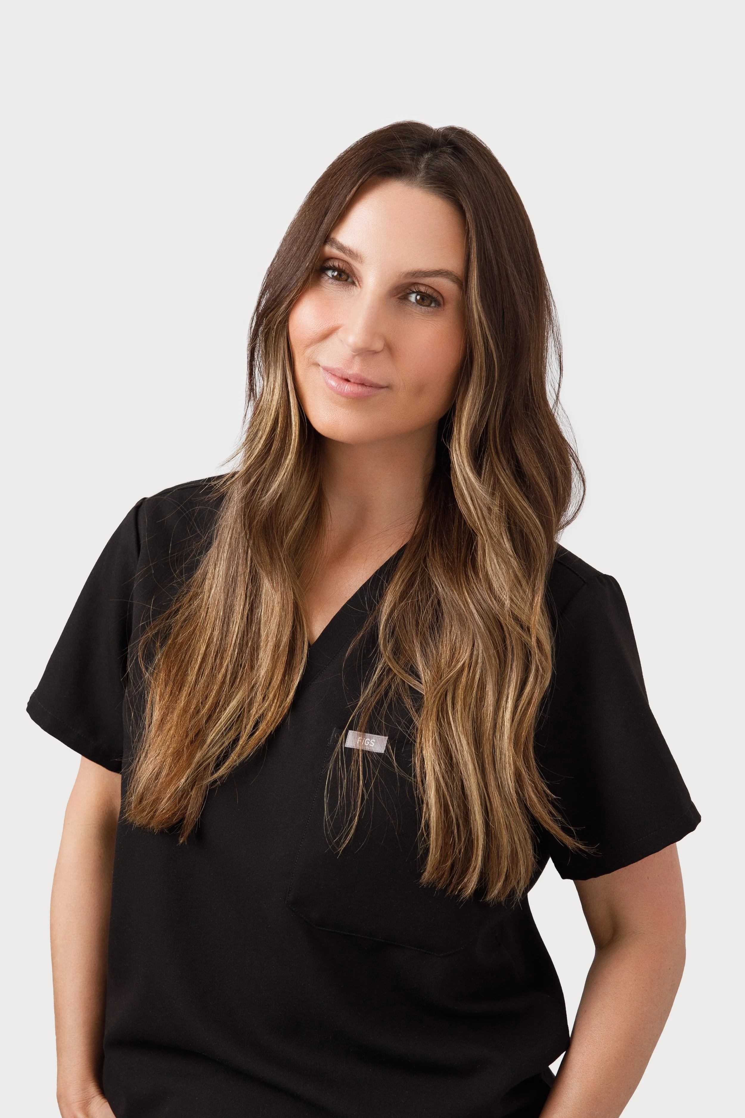 A woman with long, wavy brown hair wearing a black medical scrub top, smiling gently, against a plain white background.