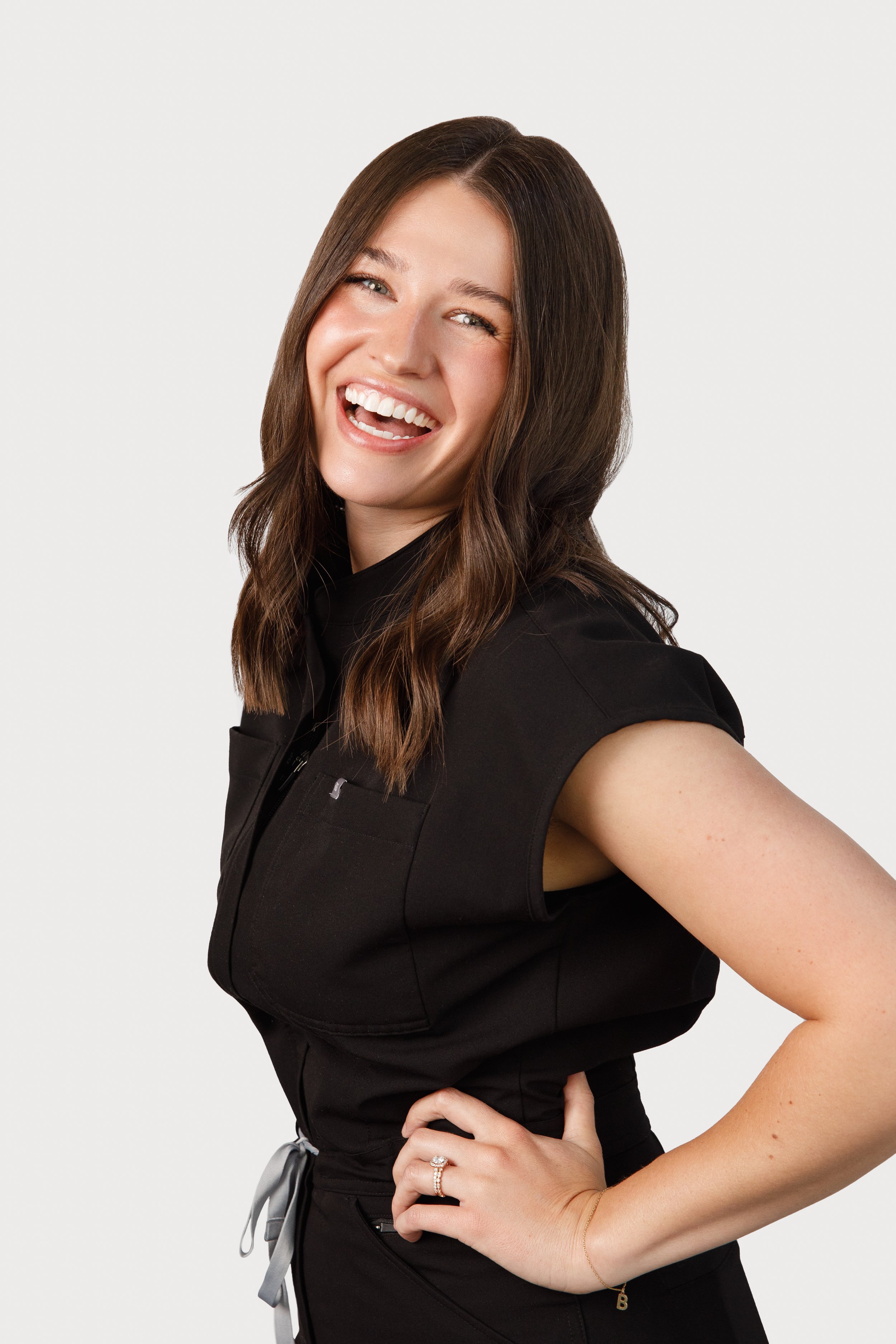 Young woman with shoulder-length brown hair wearing a sleeveless black jumpsuit, smiling, standing against a plain white background.