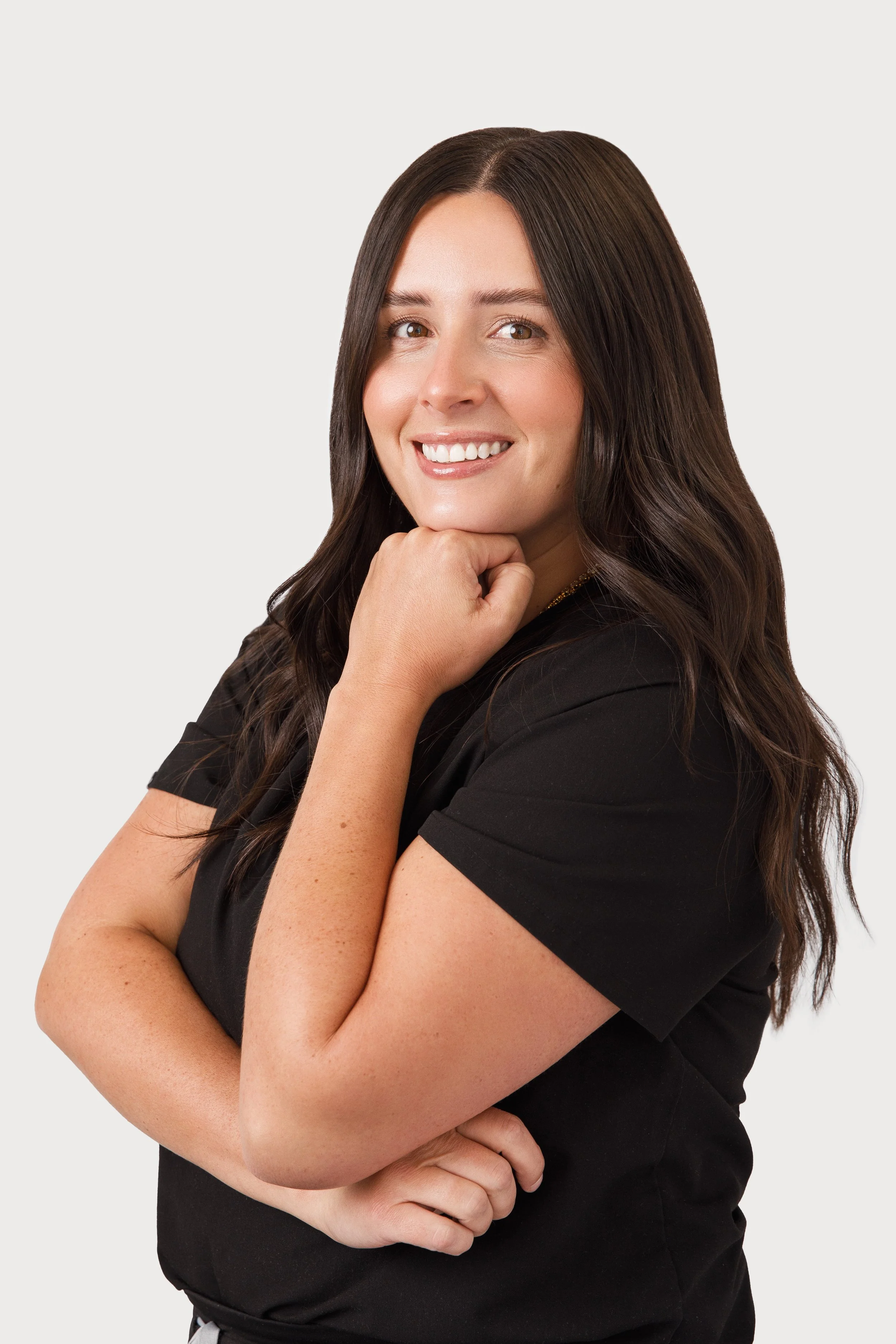 A woman with long wavy brown hair, wearing a black shirt, smiling and resting her chin on her right hand with her left arm crossed.