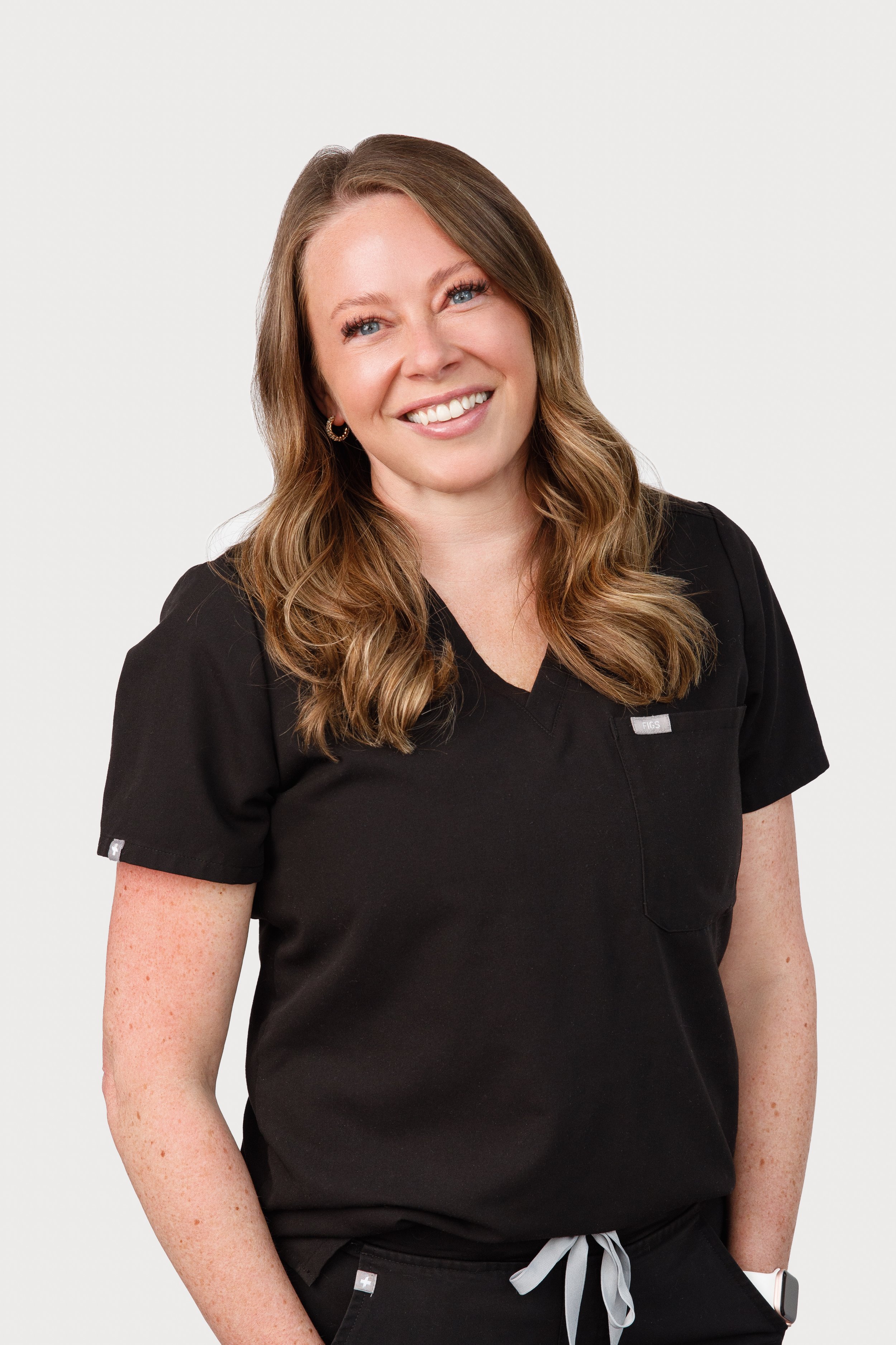 A smiling woman with wavy, light brown hair wearing black medical scrubs against a white background.