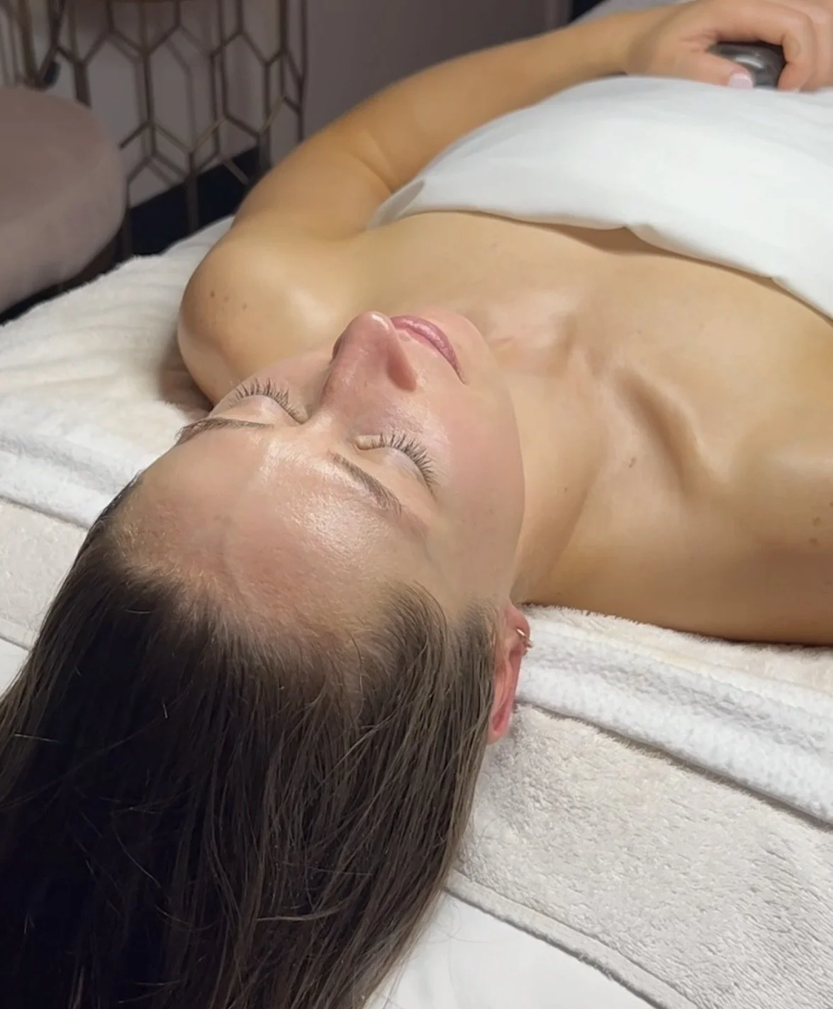A woman lying on a treatment bed with eyes closed, receiving a facial treatment, with a towel draped over her chest.