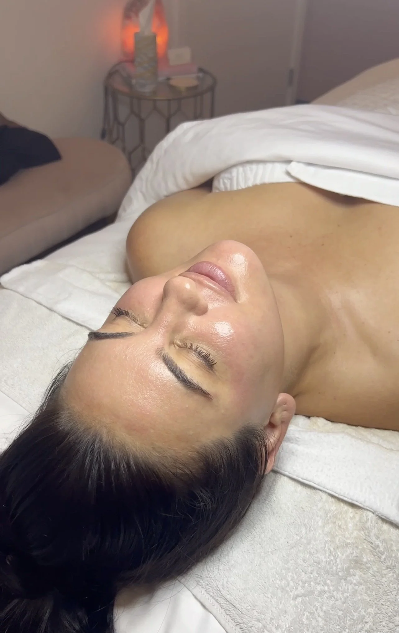 A woman lying on a treatment bed with eyes closed, receiving a facial or skincare treatment in a spa or clinic setting.