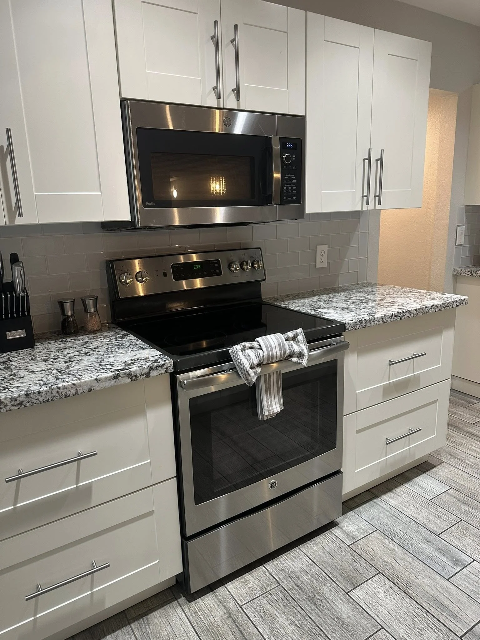 Modern kitchen with white cabinets, stainless steel microwave and oven, granite countertop, gray backsplash tiles, and wooden flooring.