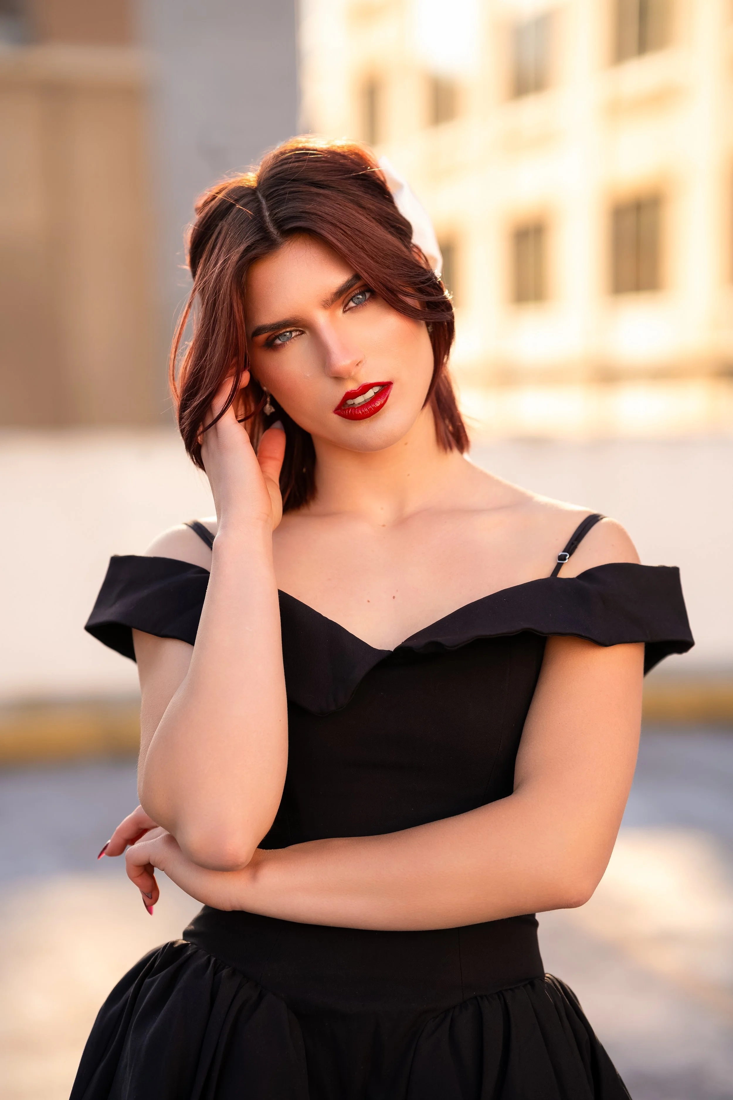 A young woman with short brunette hair and blue eyes wearing a black off-shoulder top and a black skirt standing outdoors during sunset, crossing her arms and touching her face with her right hand in front of a blurred background of buildings.