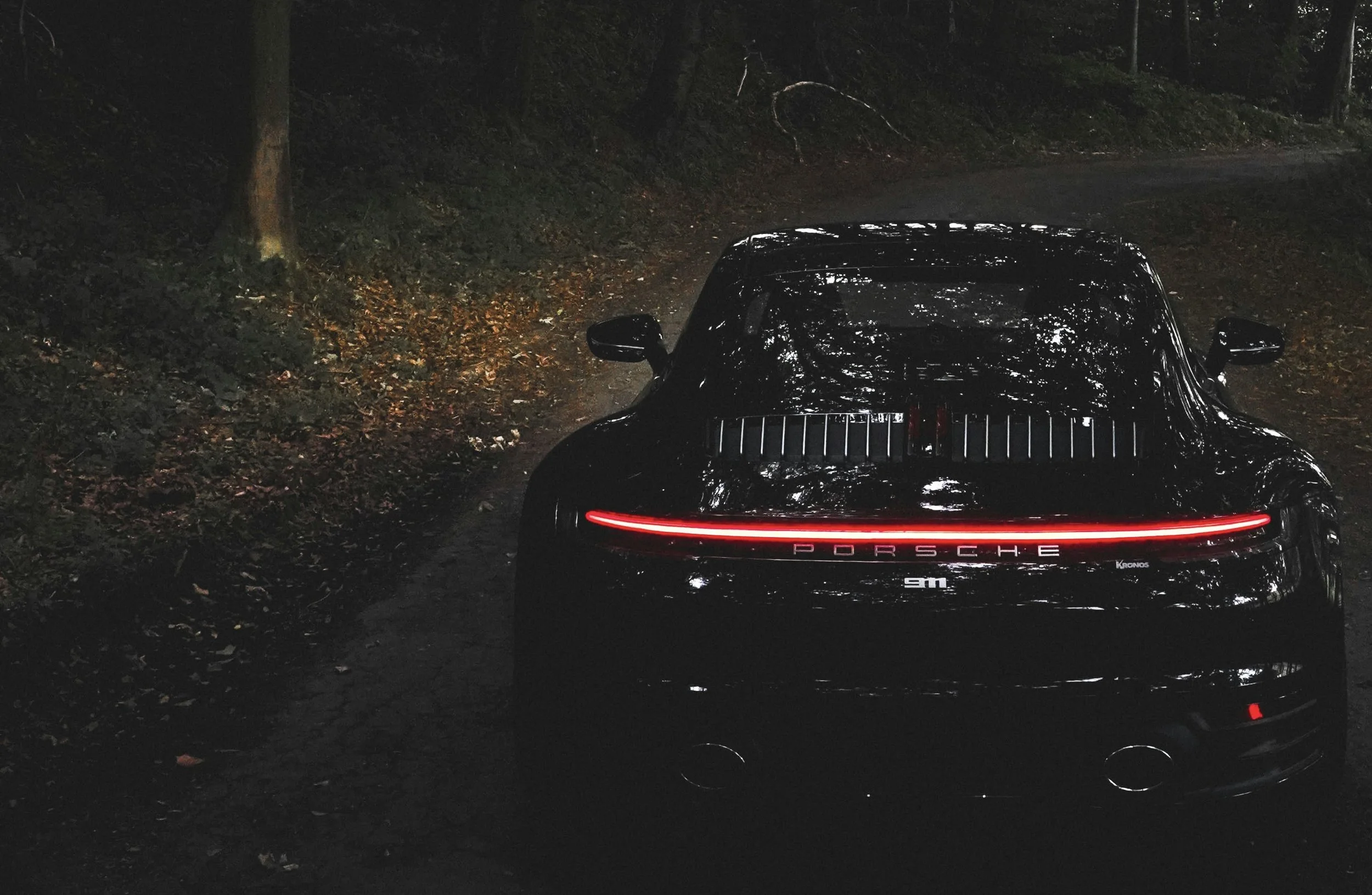 Black Porsche sports car with red rear light strip parked on a wooded forest road at night.