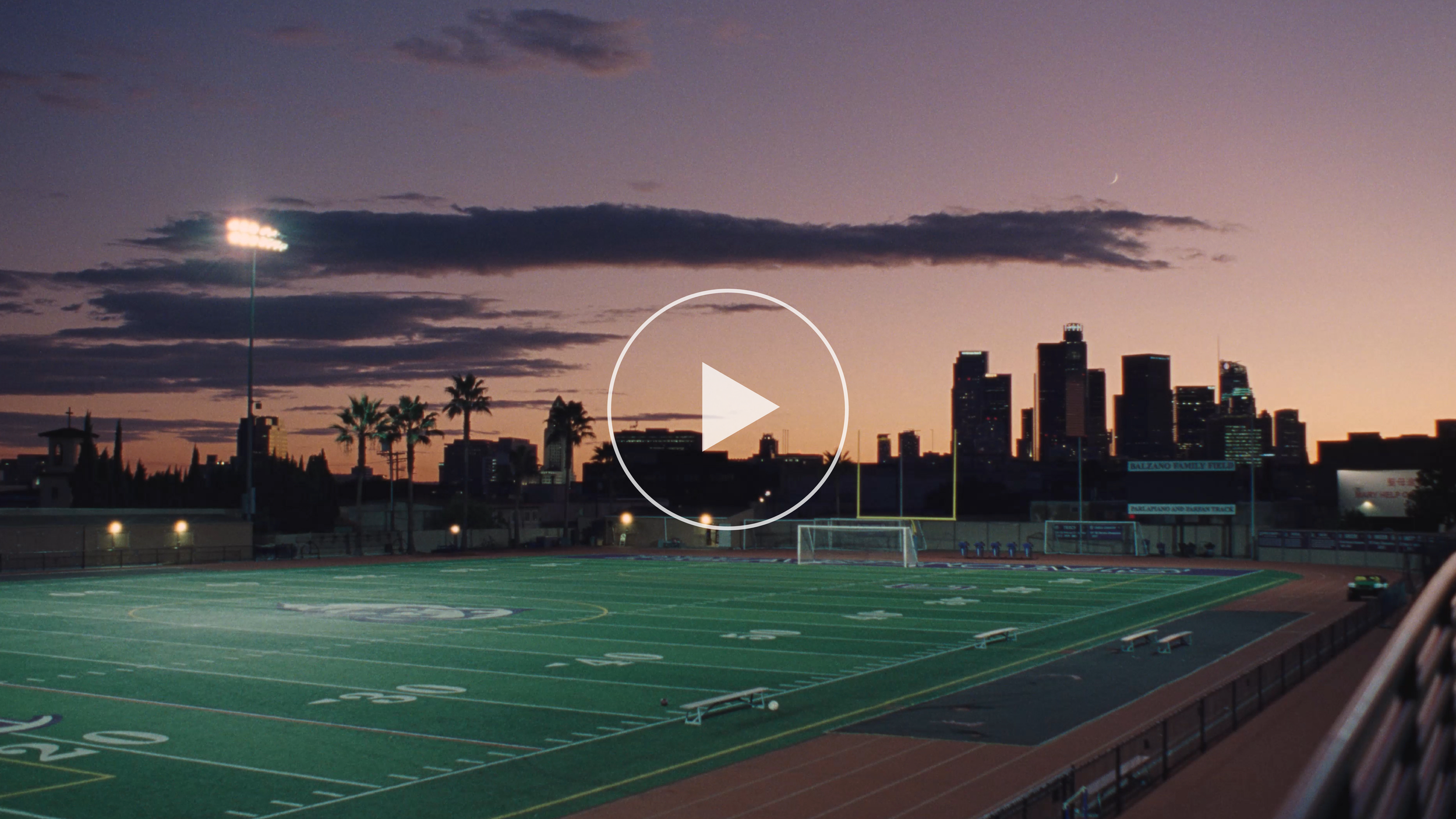 An outdoor football field with a city skyline in the background during sunset, featuring tall buildings and cloudy sky.