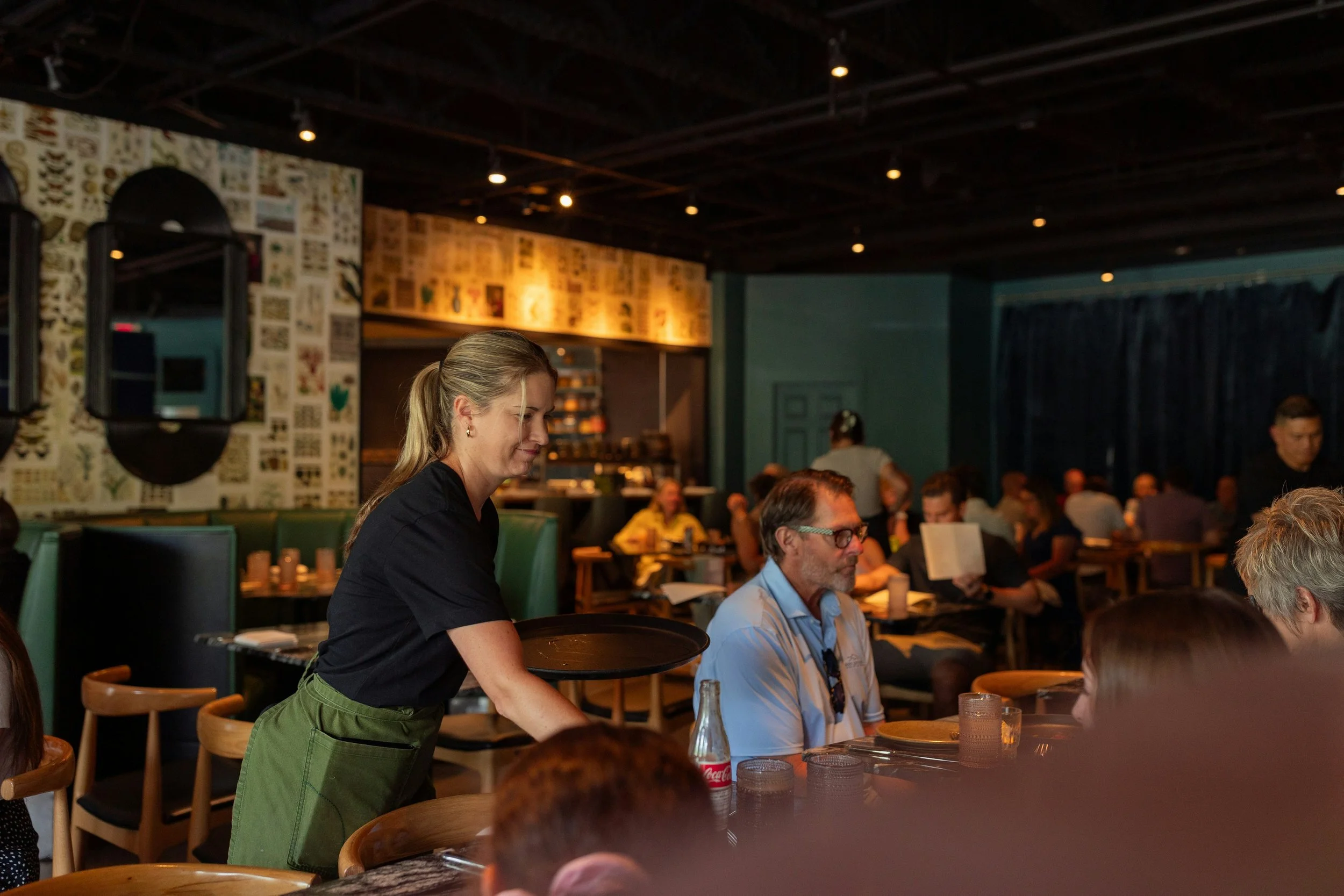 A waitress serving drinks to customers in a crowded restaurant with wood accents and wall art.