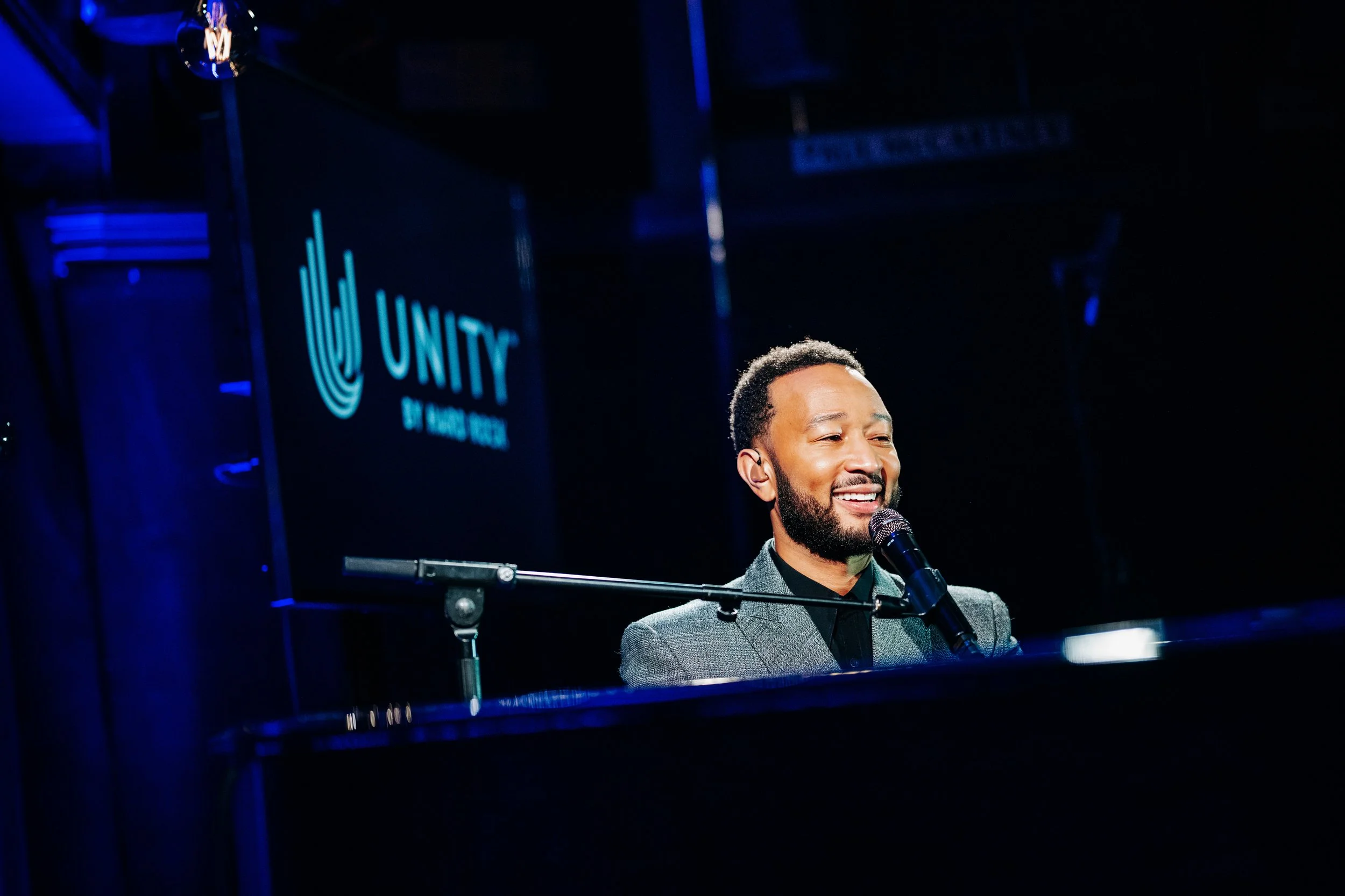 Twelve-time Grammy winner John Legend smiling while performing live at the piano for an exclusive Unity event at the Hard Rock Cafe in London. High-profile celebrity showcase and premium corporate music event photography by Will Bailey.