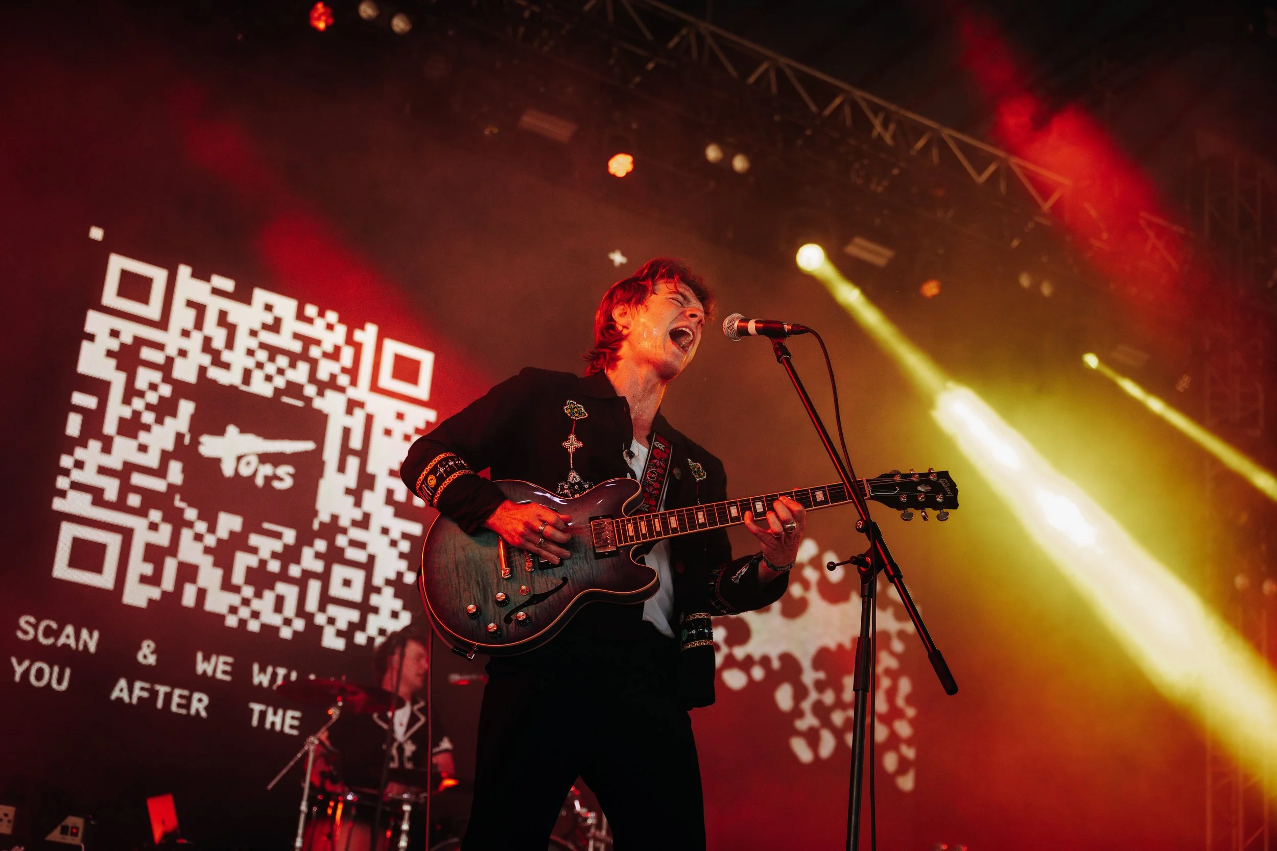 The lead singer of indie band Tors performing with his semi-hollow body electric guitar in front of a giant digital screen at Boardmasters Festival in Cornwall. Dynamic festival stage action and indie music performance photography by Will Bailey.