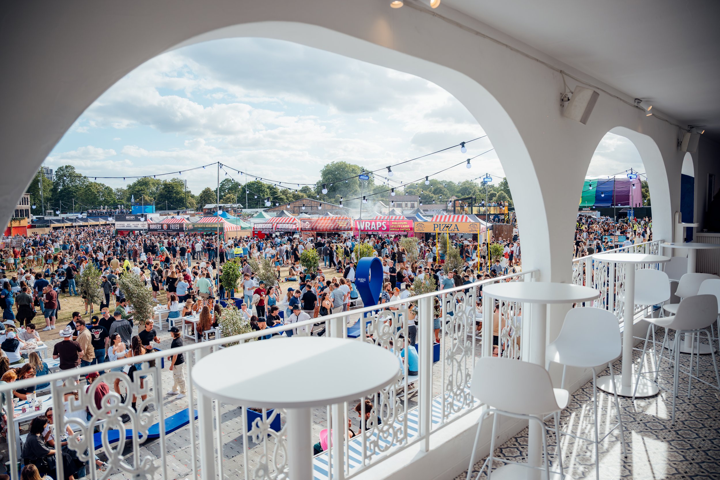 An elevated view overlooking the bustling food trucks, vendor stalls, and crowds from the upper deck of a custom brand pavilion at BST Hyde Park, London. Large-scale public festival logistics and brand infrastructure photography by Will Bailey.