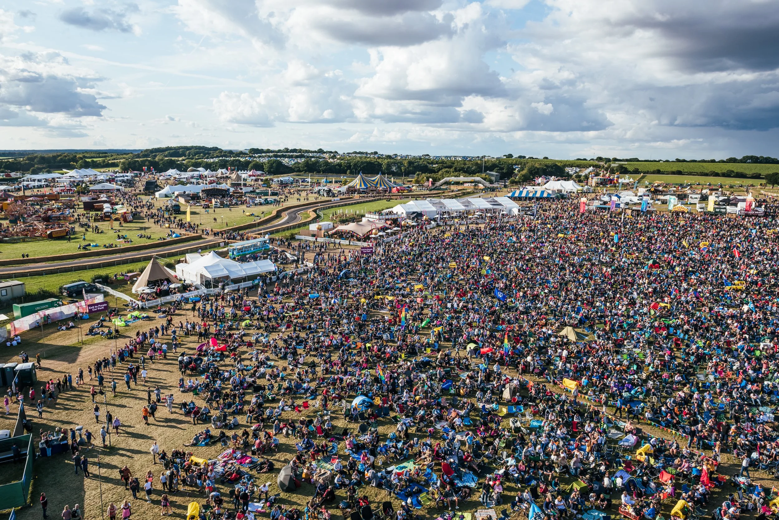 A sprawling wide aerial landscape view showcasing thousands of fans gathered across the rolling fields and main stage arena at CarFest. Large-scale music festival infrastructure and public event crowd photography by Will Bailey.
