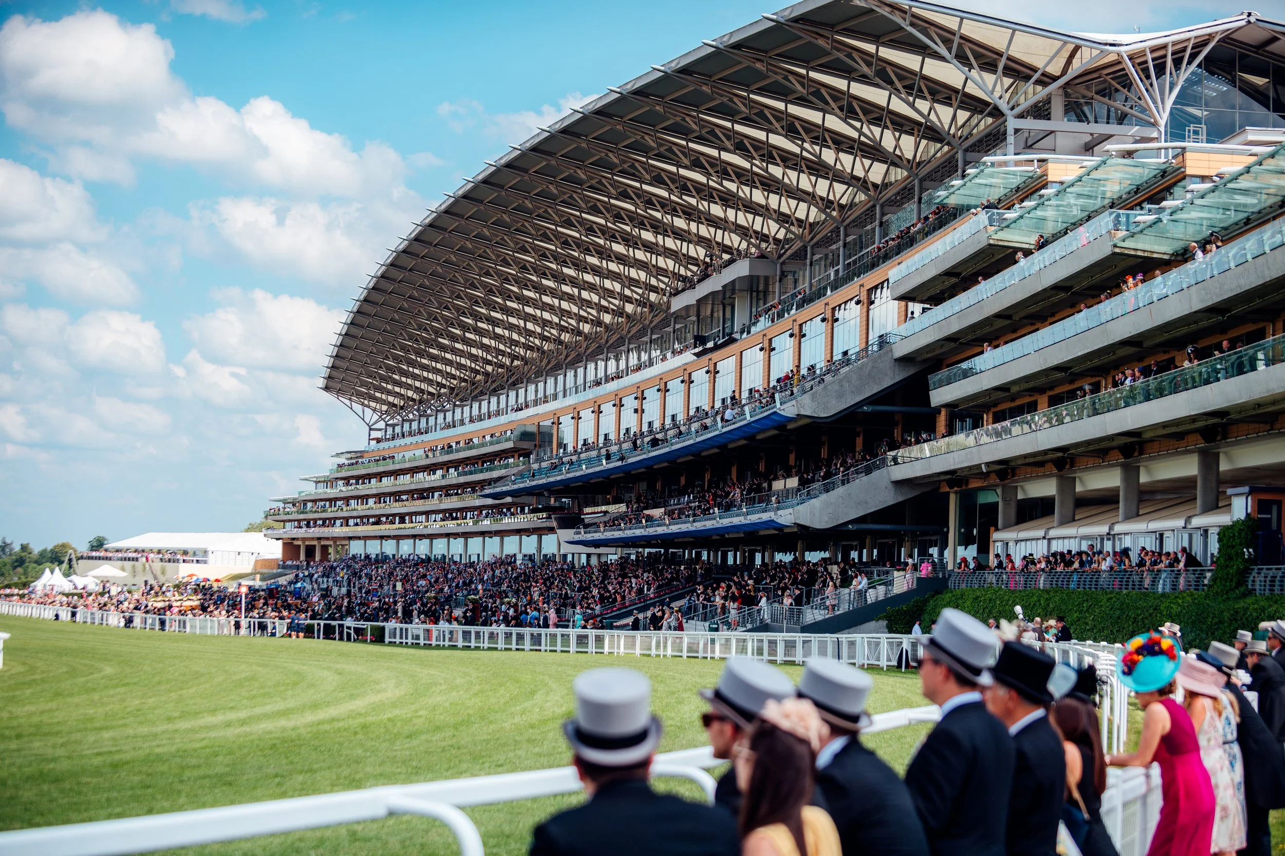 Racegoers lined up along the white rail looking out across the turf toward the iconic grandstand architecture at Royal Ascot. Elite British summer sporting season and event photography by Will Bailey.