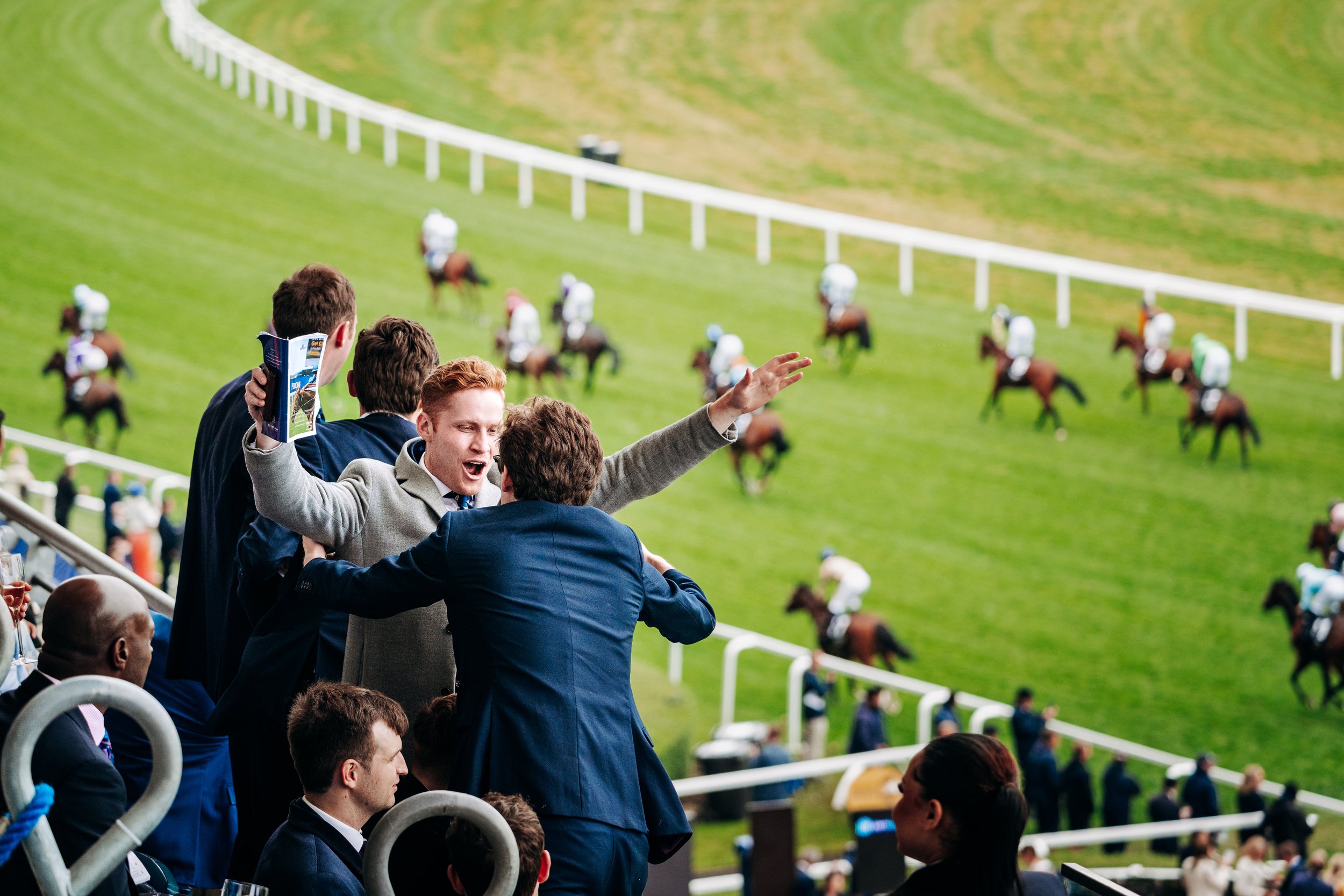 Ecstatic racegoers cheering and embracing in the grandstand as their horse crosses the finish line at Ascot Racecourse, Berkshire. Candid sports emotion and summer social season lifestyle photography by Will Bailey.