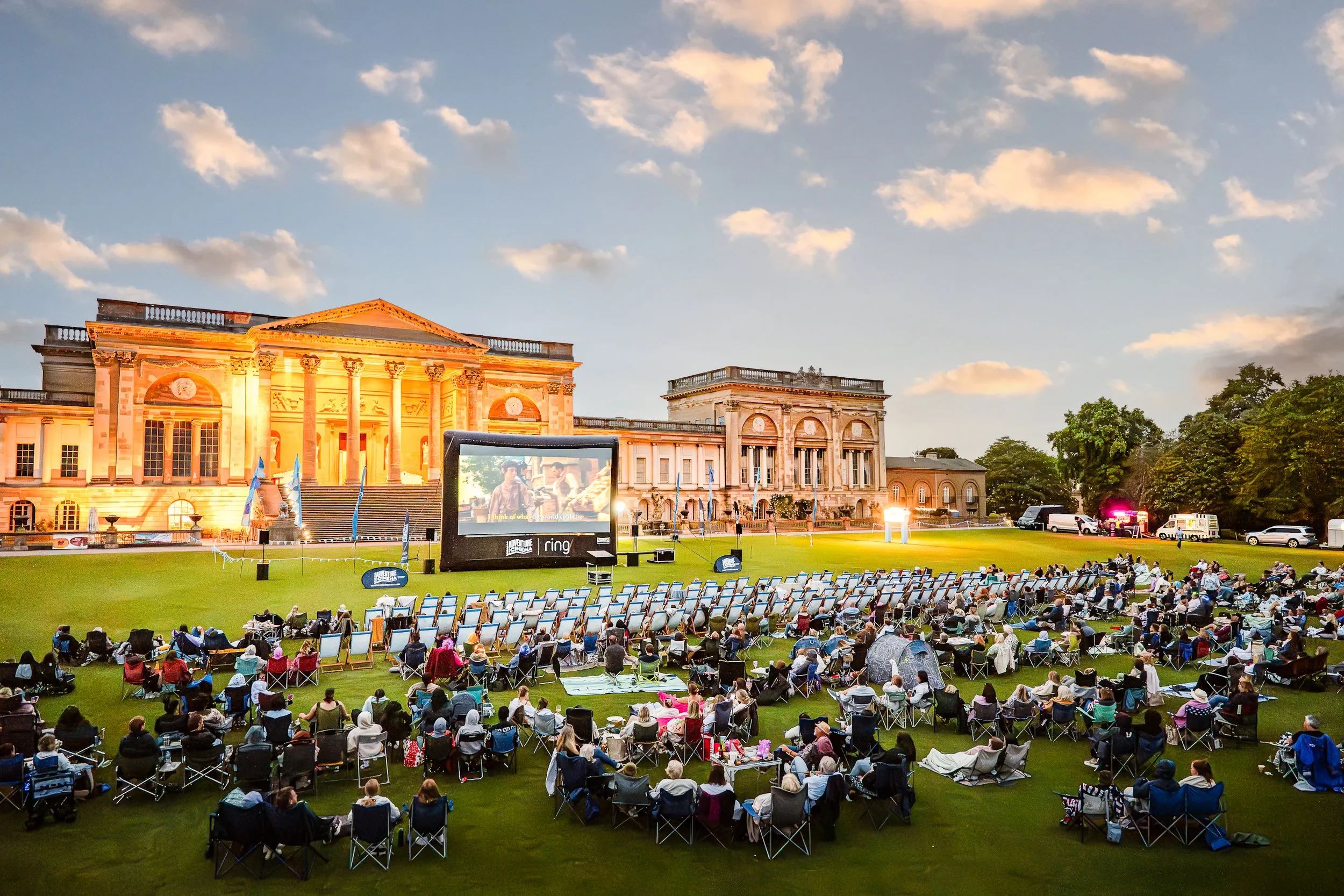 An expansive outdoor audience watching a film on a giant inflatable screen on the lawn of Stowe House during an Adventure Cinema sunset screening. Public experiential cinema and heritage venue event photography by Will Bailey.