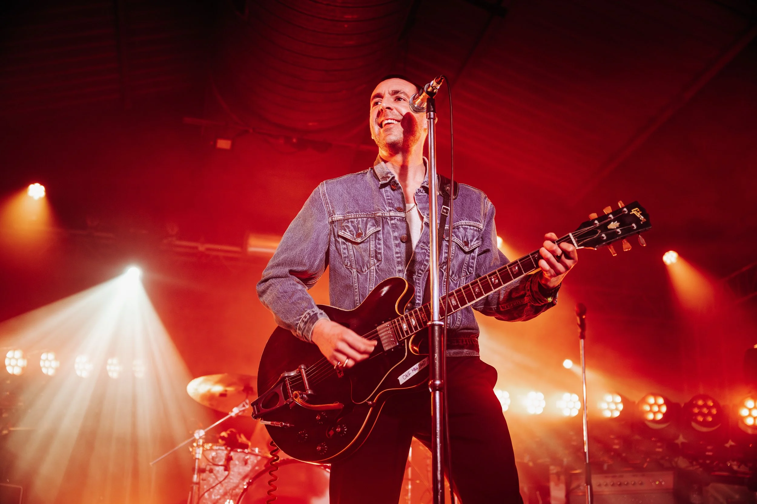 Miles Kane smiling on stage mid-song while playing his semi-hollow body electric guitar under warm lights at a live venue in Southampton, Hampshire. Professional rock concert and touring artist lifestyle photography by Will Bailey.