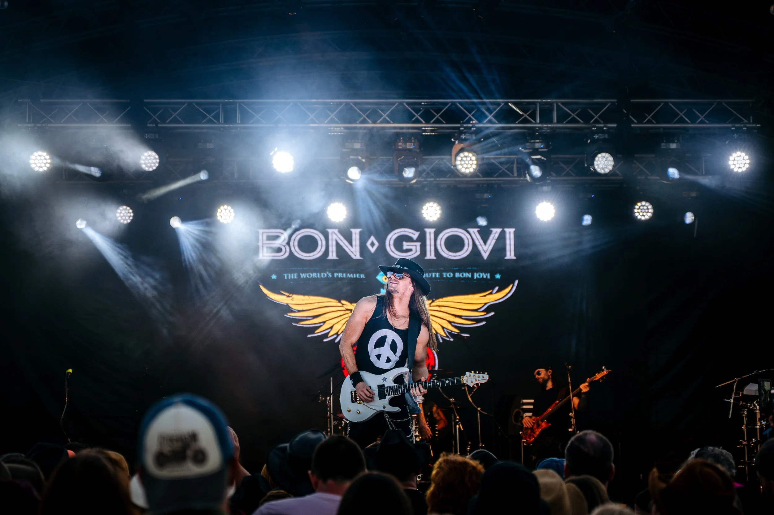 A frontman rock star pose under blinding white stage lights during a headline tribute performance by Bon Giovi at the Rock n Ribs Festival. Large-scale public entertainment and summer music festival photography by Will Bailey.