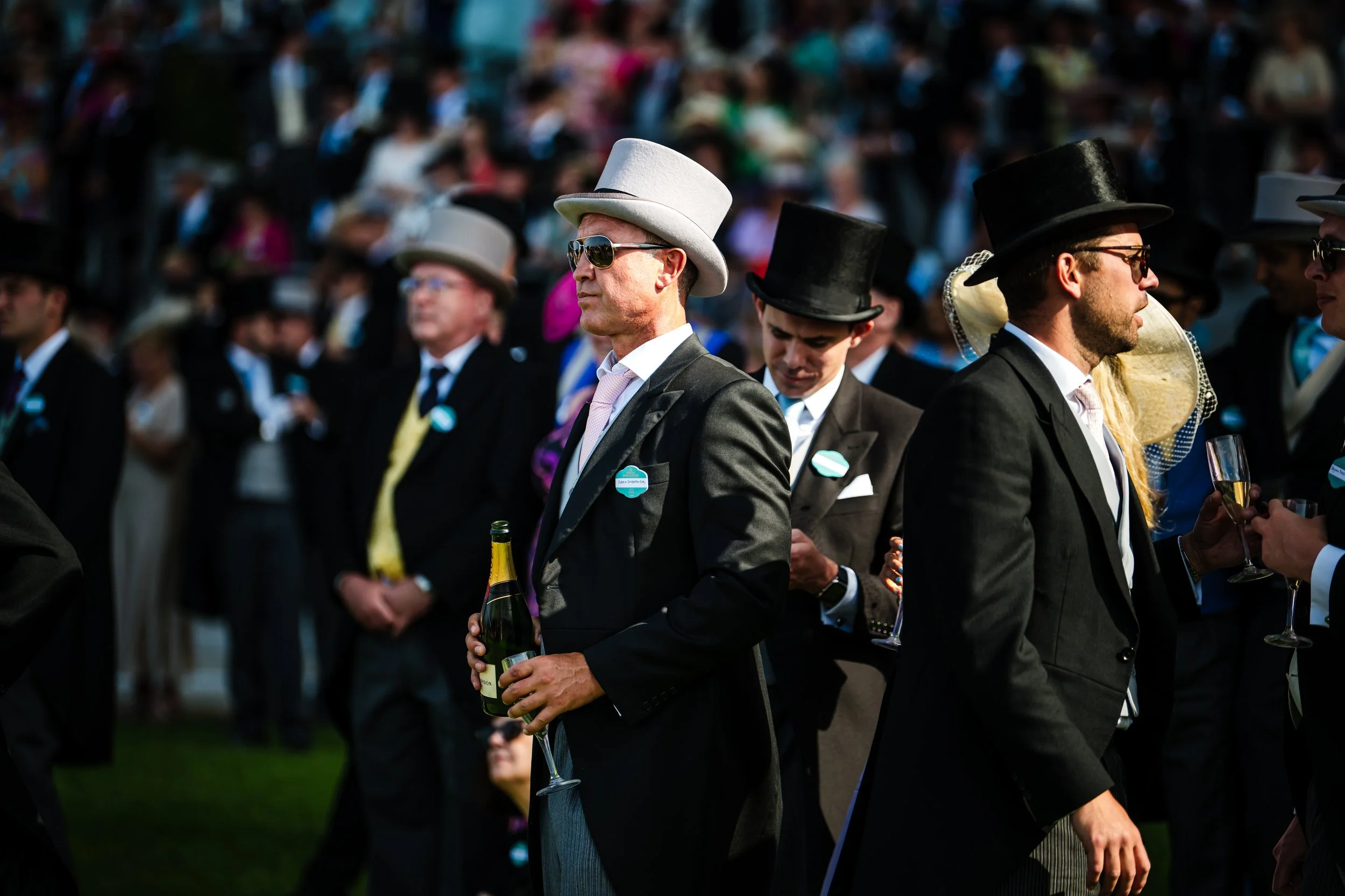 Guests in top hats and tailored morning suits enjoying champagne on the lawn during the summer social season at Royal Ascot, Berkshire. Luxury hospitality and high-end lifestyle event photography by Will Bailey.