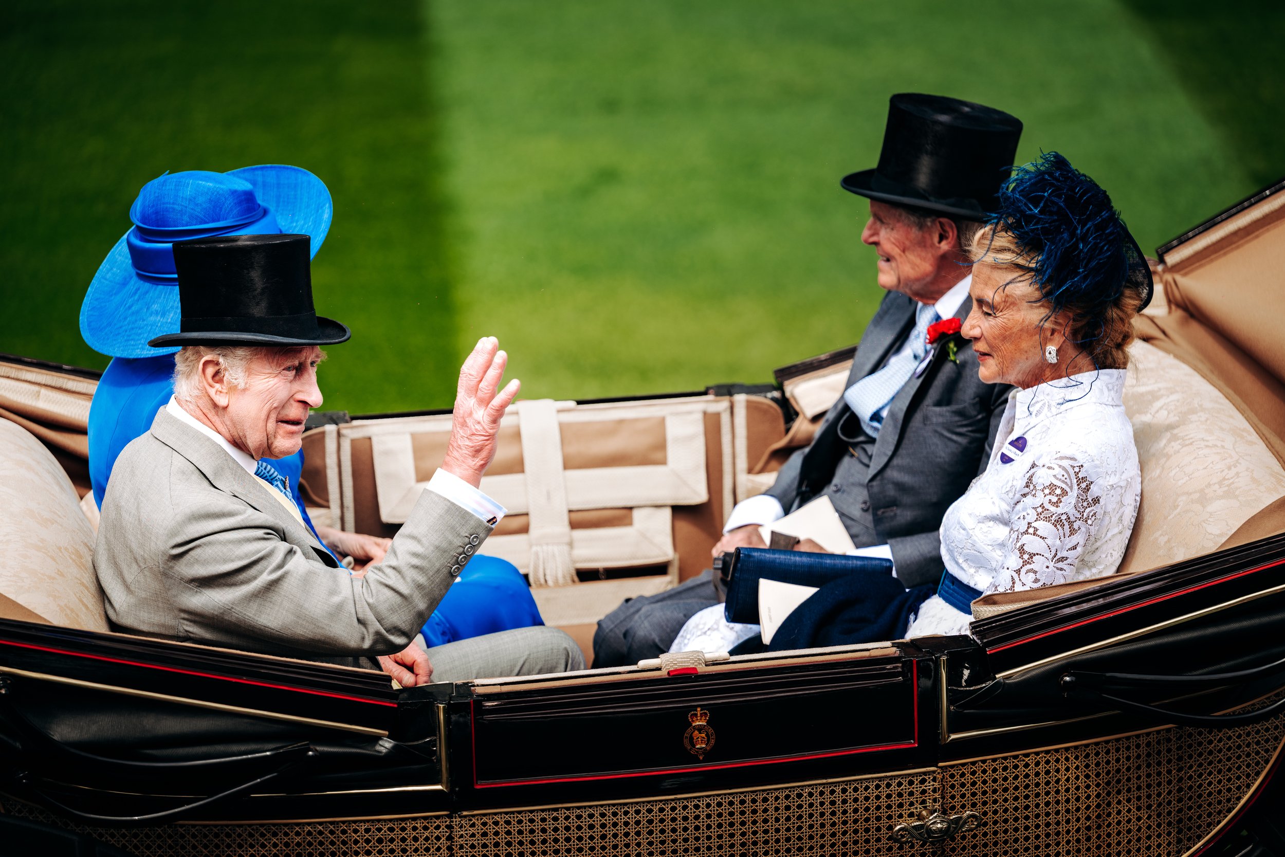 King Charles III waving to spectators from an open horse-drawn carriage during the traditional Royal Procession arrival at Royal Ascot, Berkshire. Editorial documentary and high-profile societal event photography by Will Bailey.