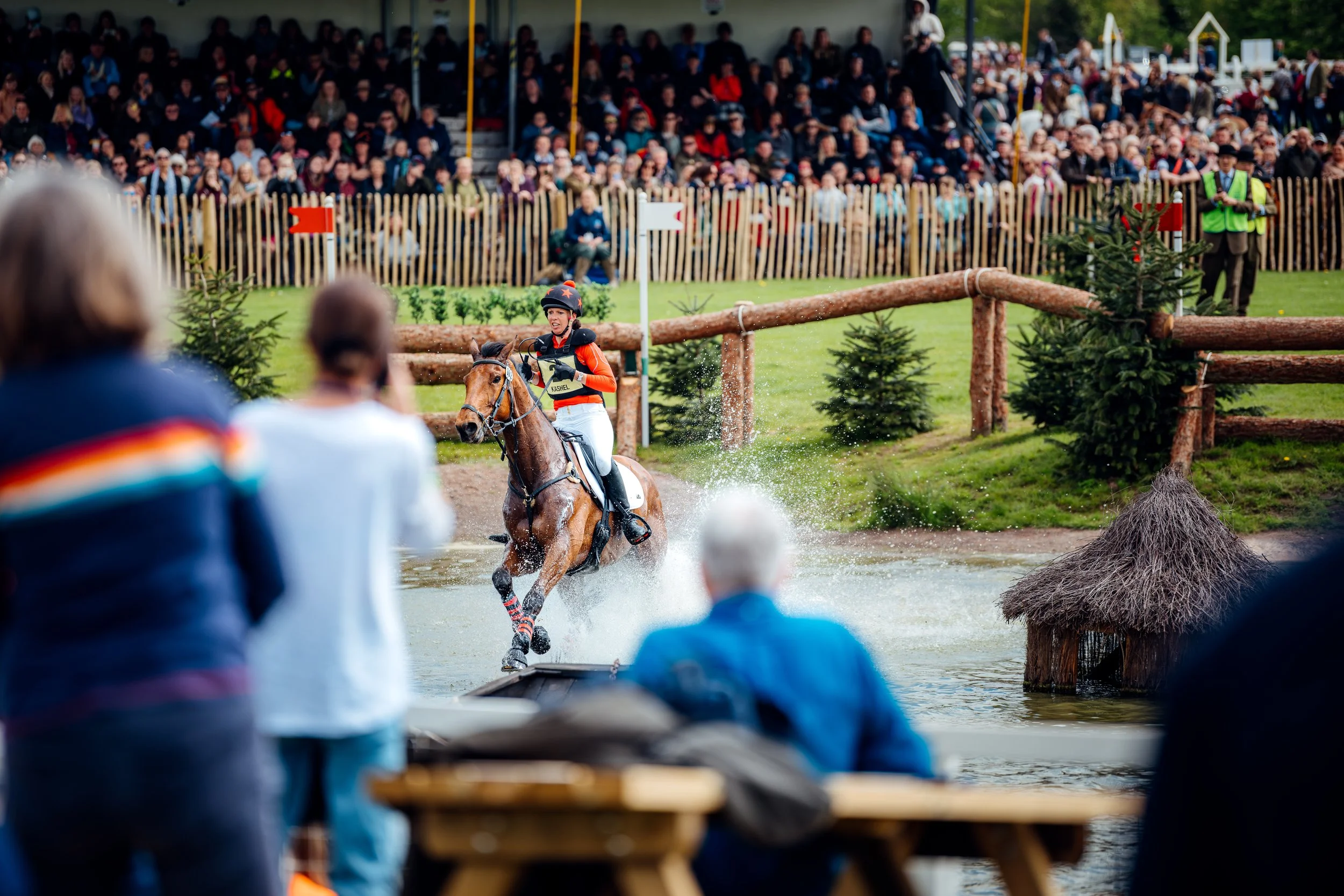 An equestrian competitor splashing through the water complex in front of spectators during the cross-country phase of the Badminton Horse Trials in Gloucestershire. Elite eventing and Cotswolds country lifestyle estate sports photography by Will Bail