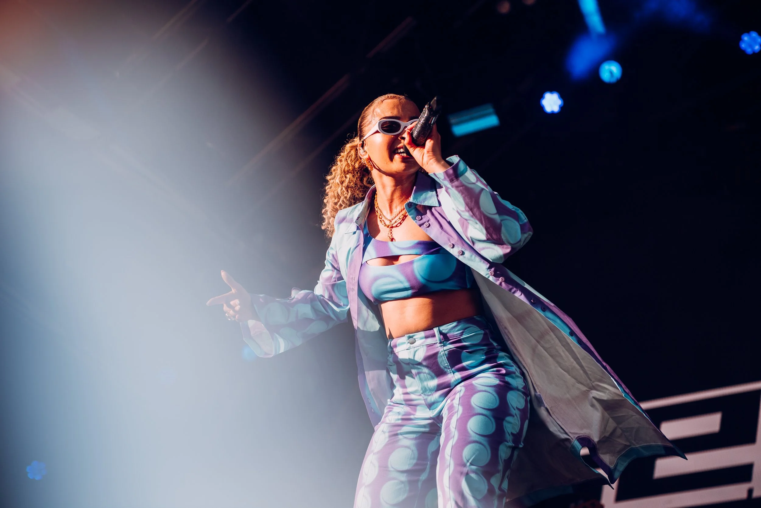 R&B singer Ella Eyre singing live in a patterned outfit and sunglasses during an energetic stage set at NASS Festival. Action-focused music and extreme sports lifestyle festival photography by Will Bailey.